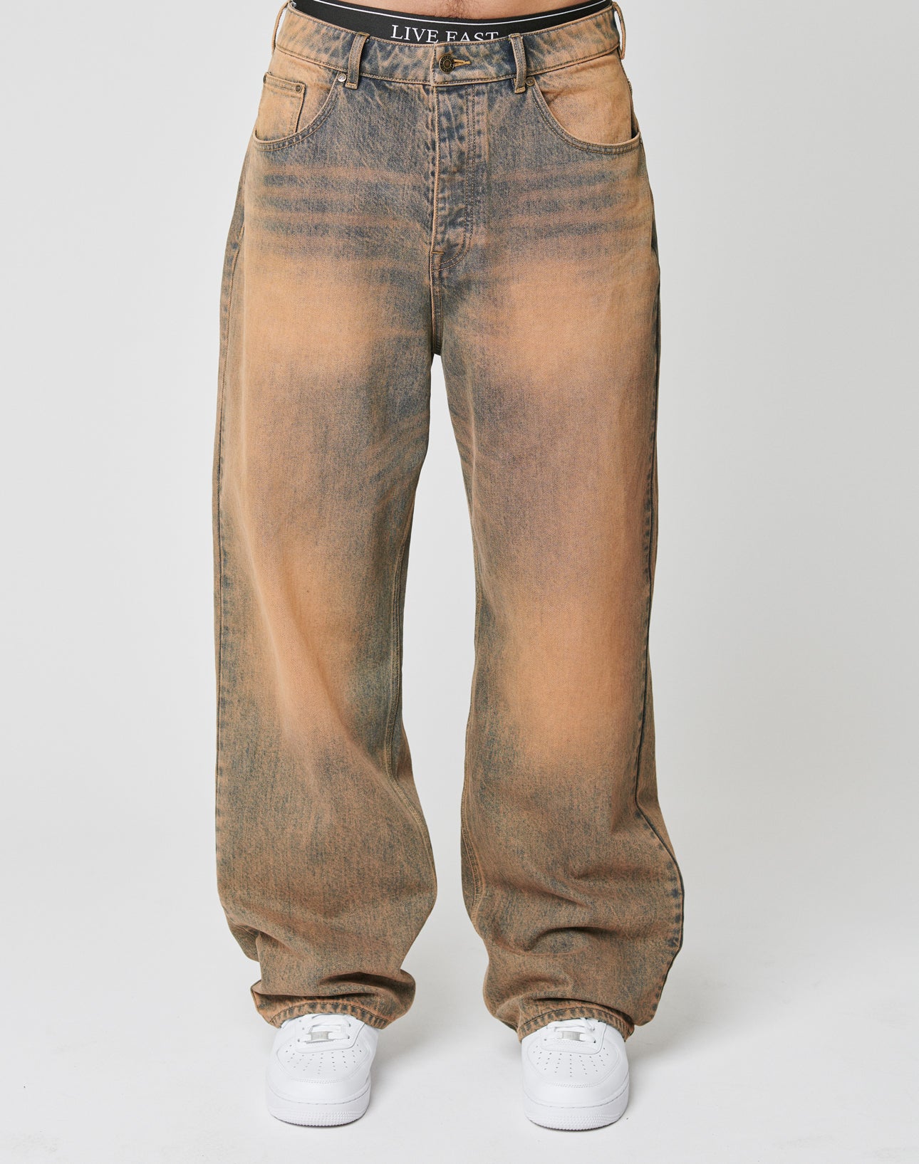 A person wears LFDY Baggy Denim by LIVE FAST DIE YOUNGābaggy, high-waisted jeans with a faded brown-black wash and wide straight leg. Paired with white sneakers and a black waistband featuring LIVE FAST. Plain white background.