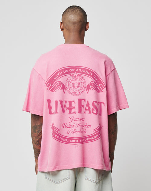 A person wears the LFDY Loyal Tee by LIVE FAST DIE YOUNG, an oversized loose pink 100% Baumwolle shirt with a bold LIVE FAST back print featuring Germany, United Kingdom, Netherlands in decorative text and graphics.