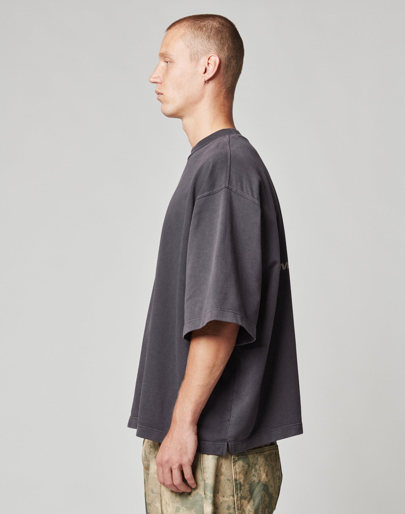 A man with short hair stands in profile against a plain background, wearing the LIVE FAST DIE YOUNG Live Fast Cropped Oversized Tee and light camouflage-patterned pants.