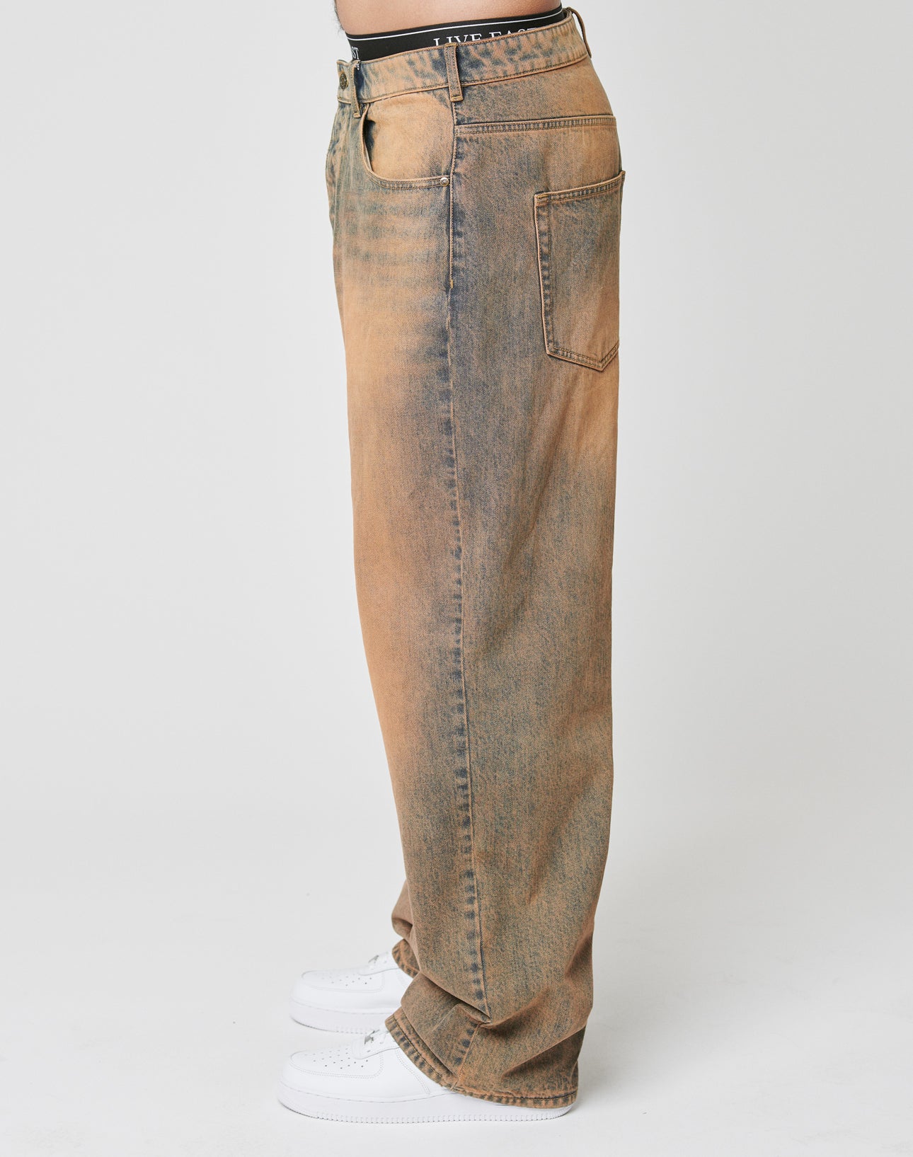 A person in profile wears LFDY Baggy Denim jeans by LIVE FAST DIE YOUNGāloose-fitting, faded brown and blue, with a baggy fit and wide straight leg. Made from 100% cotton, featuring front and back pockets, paired with white sneakers on a plain white background.