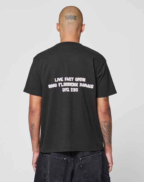 A tattooed, shaved-head individual wears the LFDY District Tee by LIVE FAST DIE YOUNG, a Regular Fit cotton shirt with front and back prints; the back reads: LIVE FAST GMBH SOHO FLAGSHIP MARAIS RCC 235. They stand before a plain background.