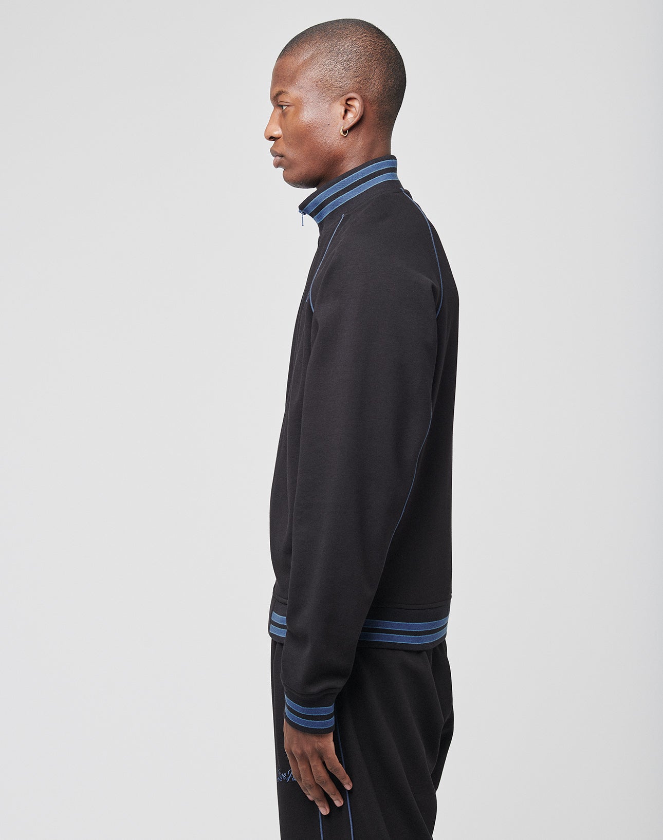 A man stands in profile against a plain light background, wearing the LIVE FAST DIE YOUNG Live Fast Tracksuit Jacket with blue trim, Raglan-Ärmel, and Ripp-Kragen, paired with matching pants for a sporty look.