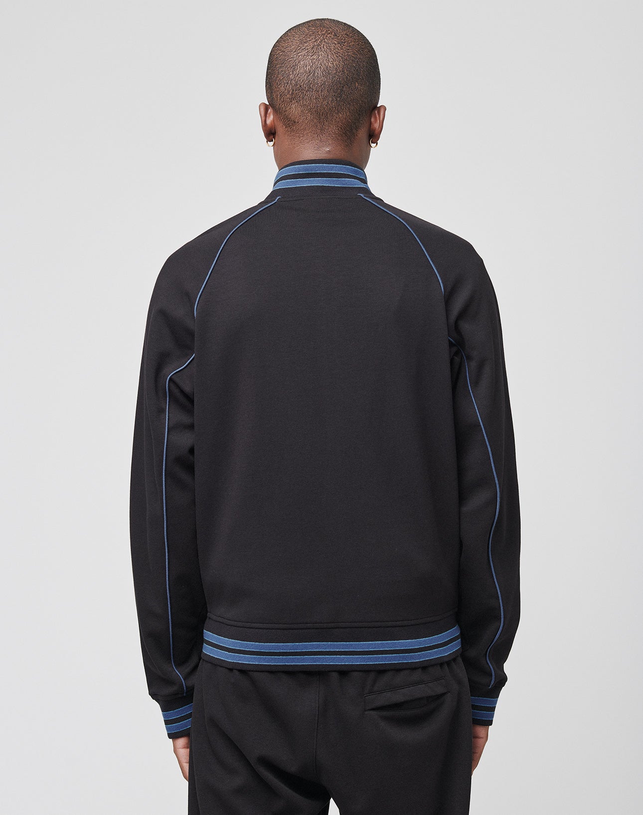 A person faces away from the camera in a black LIVE FAST DIE YOUNG Live Fast Tracksuit Jacket with blue trim, blue piping, logo embroidery on the chest, and striped ribbed collar, cuffs, and waistband. The background is plain light gray.