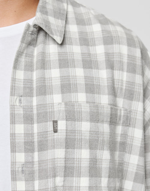 A close-up of a person in the LIVE FAST DIE YOUNG LFDY Light Flannel, a gray and white plaid oversize shirt with a chest pocket and small fabric tag, layered over a white tee; only their chest and neck are visible.