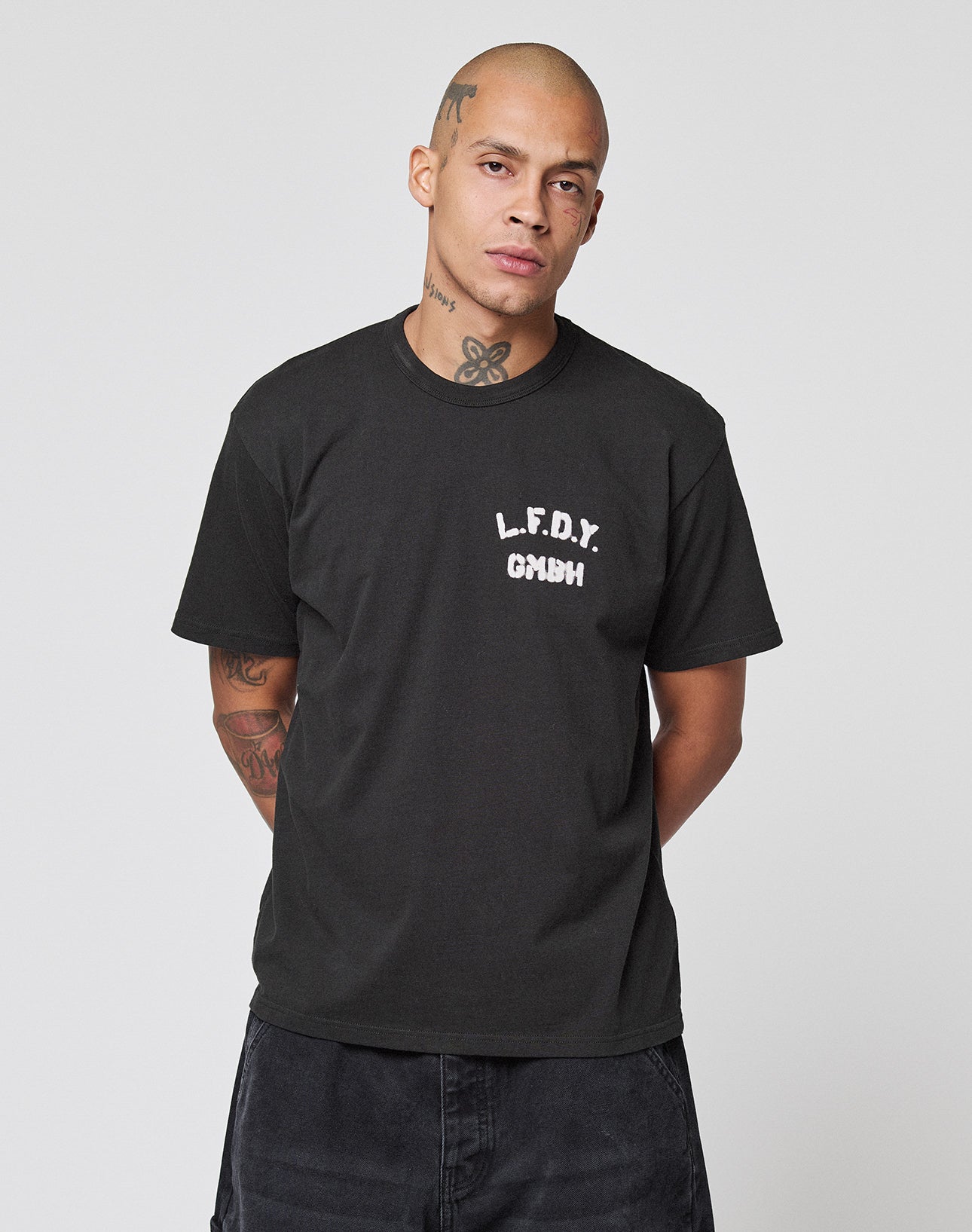 A person with tattoos on their neck and arms stands against a plain background, wearing the LIVE FAST DIE YOUNG LFDY District Tee with white L.F.D.Y. GMBH text on the left chest.