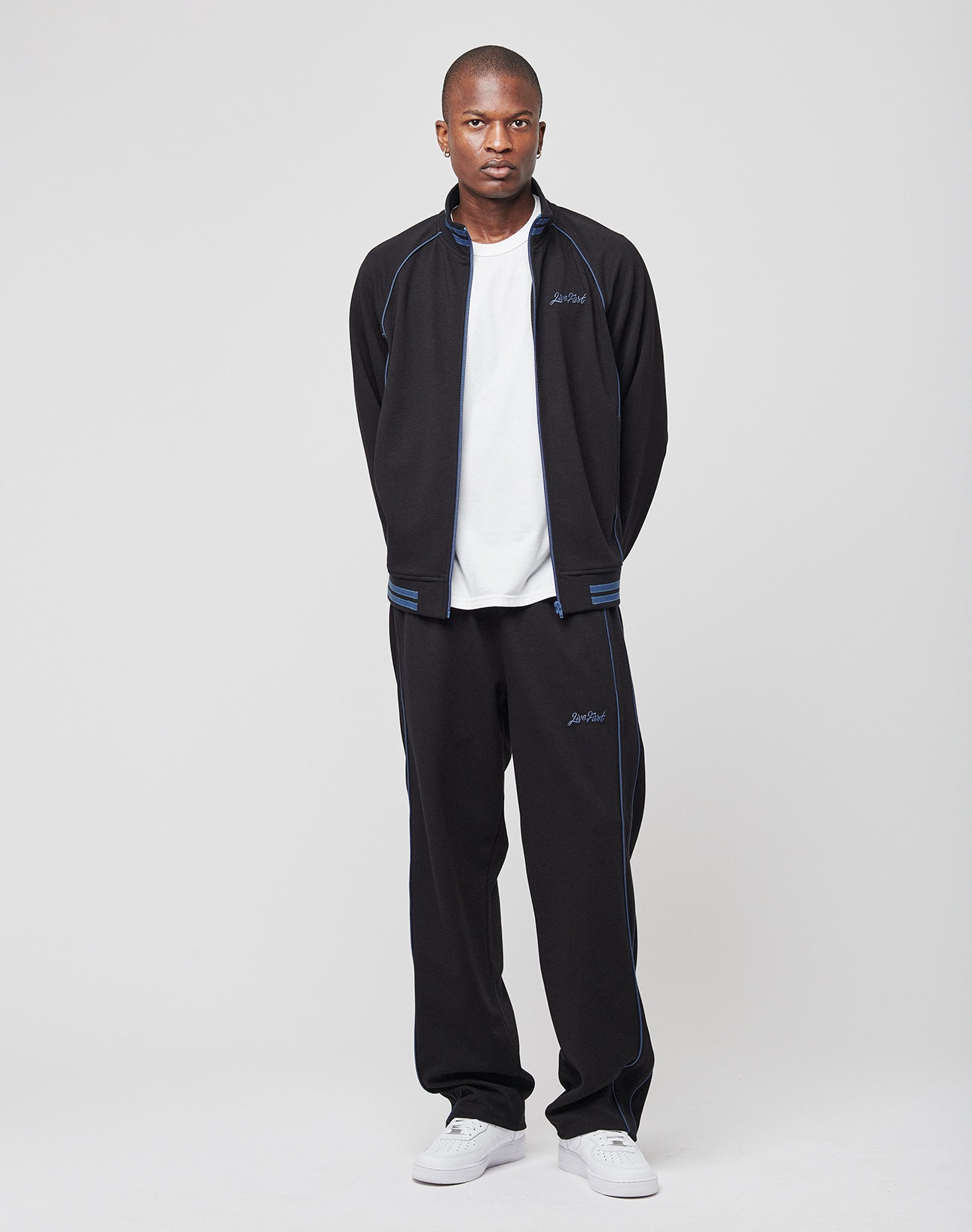 A man faces forward wearing the LIVE FAST DIE YOUNG Live Fast Tracksuit Jacket in black with blue raglan sleeves and a ribbed collar, paired with a white t-shirt and sneakers, hands behind his back, against a plain light gray background.