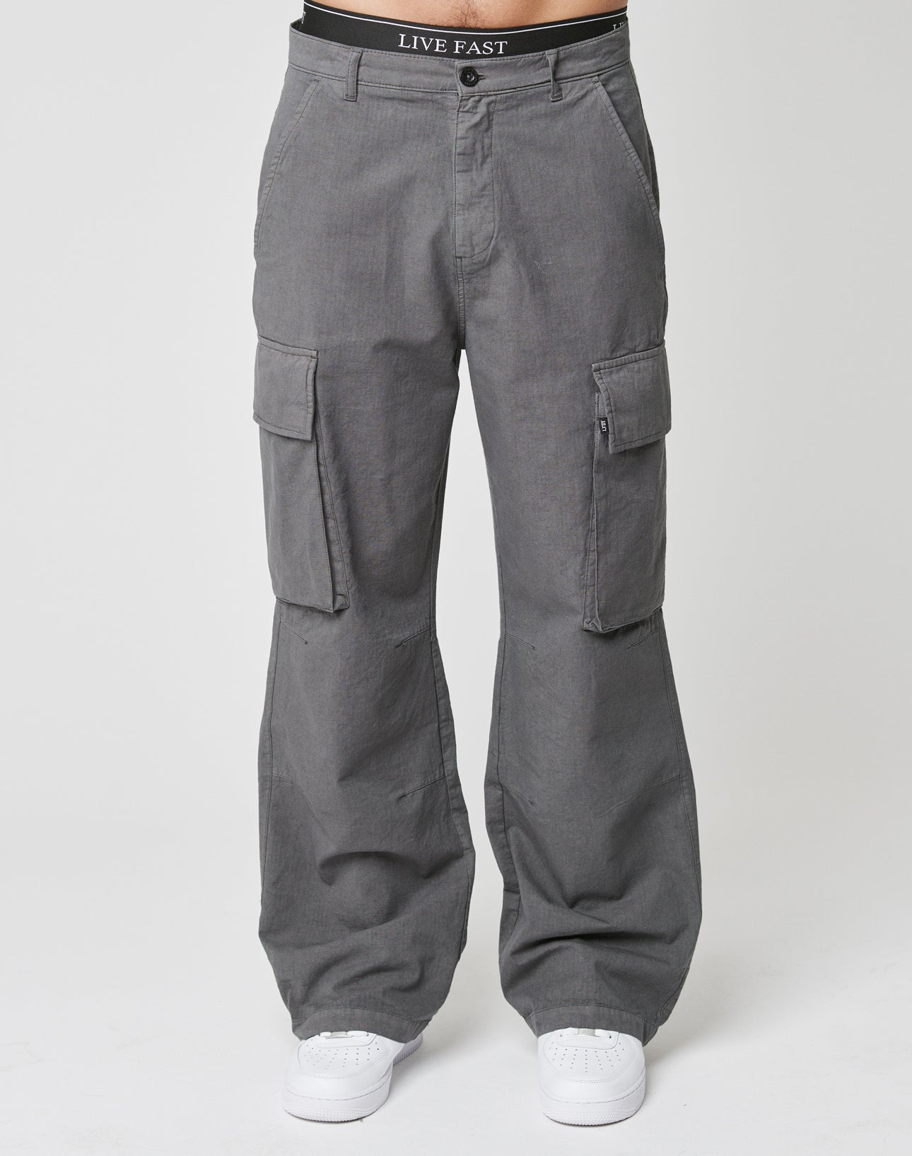 A person wears the LIVE FAST DIE YOUNG Wide Cargoāa gray, wide fit pant with Cargo-Taschen and side pocketsāstyled with white sneakers. The black waistband reads LIVE FAST. Theyre standing against a plain white background.