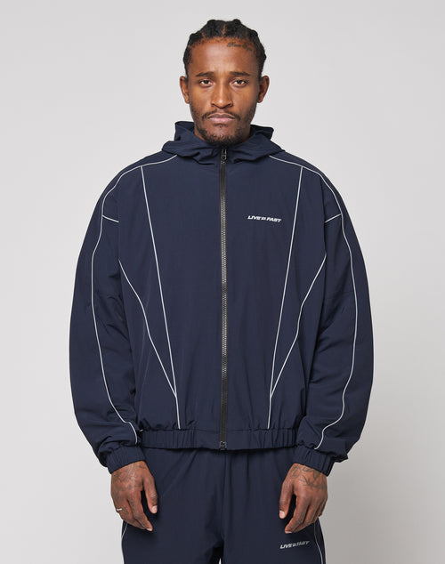 A man wears the LIVE FAST DIE YOUNG Live Fast Trackjacket in navy blue with reflektierender Druck, white piping, zip pockets, elastic cuffs, and LIVE FAST text on the chest, paired with matching pants against a light gray background.