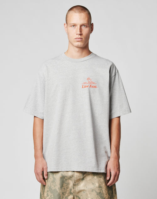 A person with short hair wears the LIVE FAST DIE YOUNG LFDY Praying Tee, an oversize light gray T-shirt with red Live Fast text, standing against a plain light gray background and wearing tan and green patterned pants.