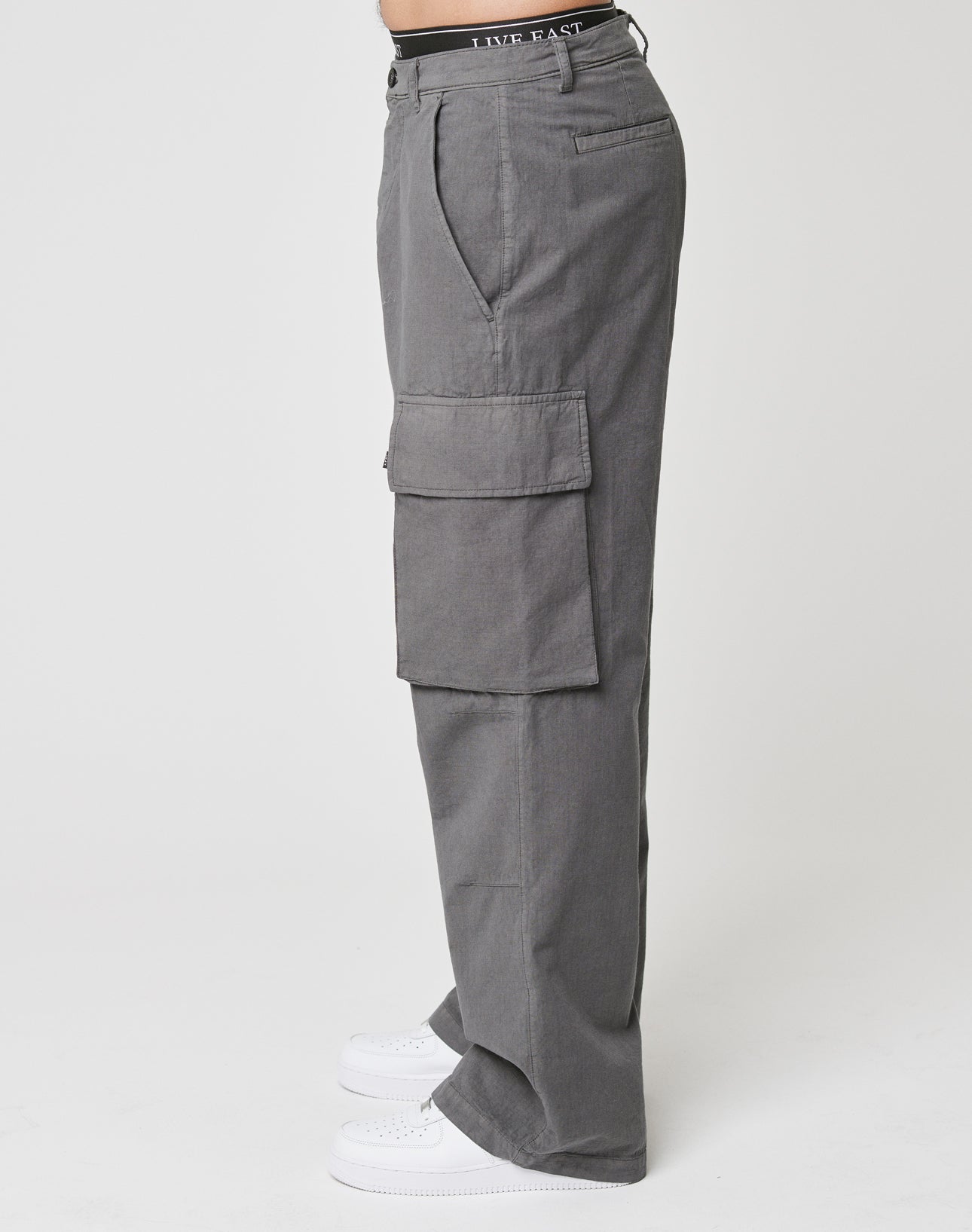 A person in LIVE FAST DIE YOUNG Wide Cargo high-waisted gray ripstop cargo pants with large pockets and a wide fit, styled with white sneakers, is shown in side profile against a plain white background.