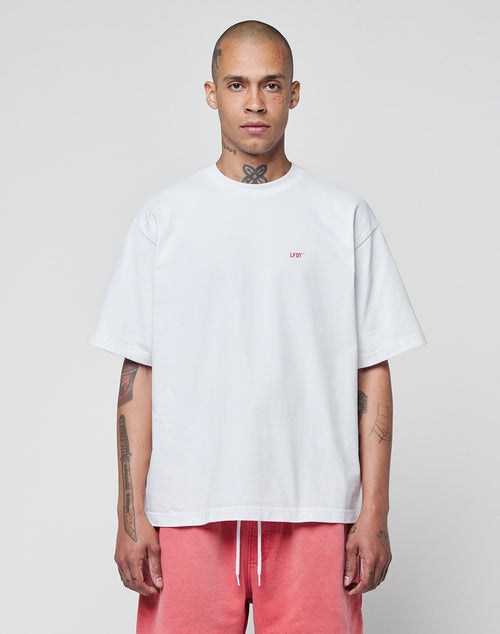 A person with a shaved head and tattoos faces forward, wearing the LIVE FAST DIE YOUNG Basic Evidence Tee in white with small red “LBY” lettering, paired with pink drawstring shorts, set against a plain light background.
