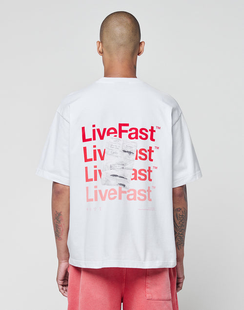 Seen from behind, a person with a shaved head wears the LIVE FAST DIE YOUNG Basic Evidence Tee—a white, 100% Baumwolle oversize T-shirt with red LiveFast prints on front and back—paired with pink shorts against a plain light background.