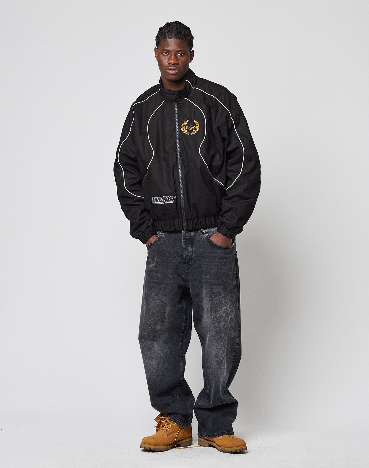 A man with short braided hair and a neutral expression stands against a plain background wearing LIVE FAST DIE YOUNG LFDY Tire Tracks Denim, a black jacket with white piping, and tan work boots, with one hand in his pocket.