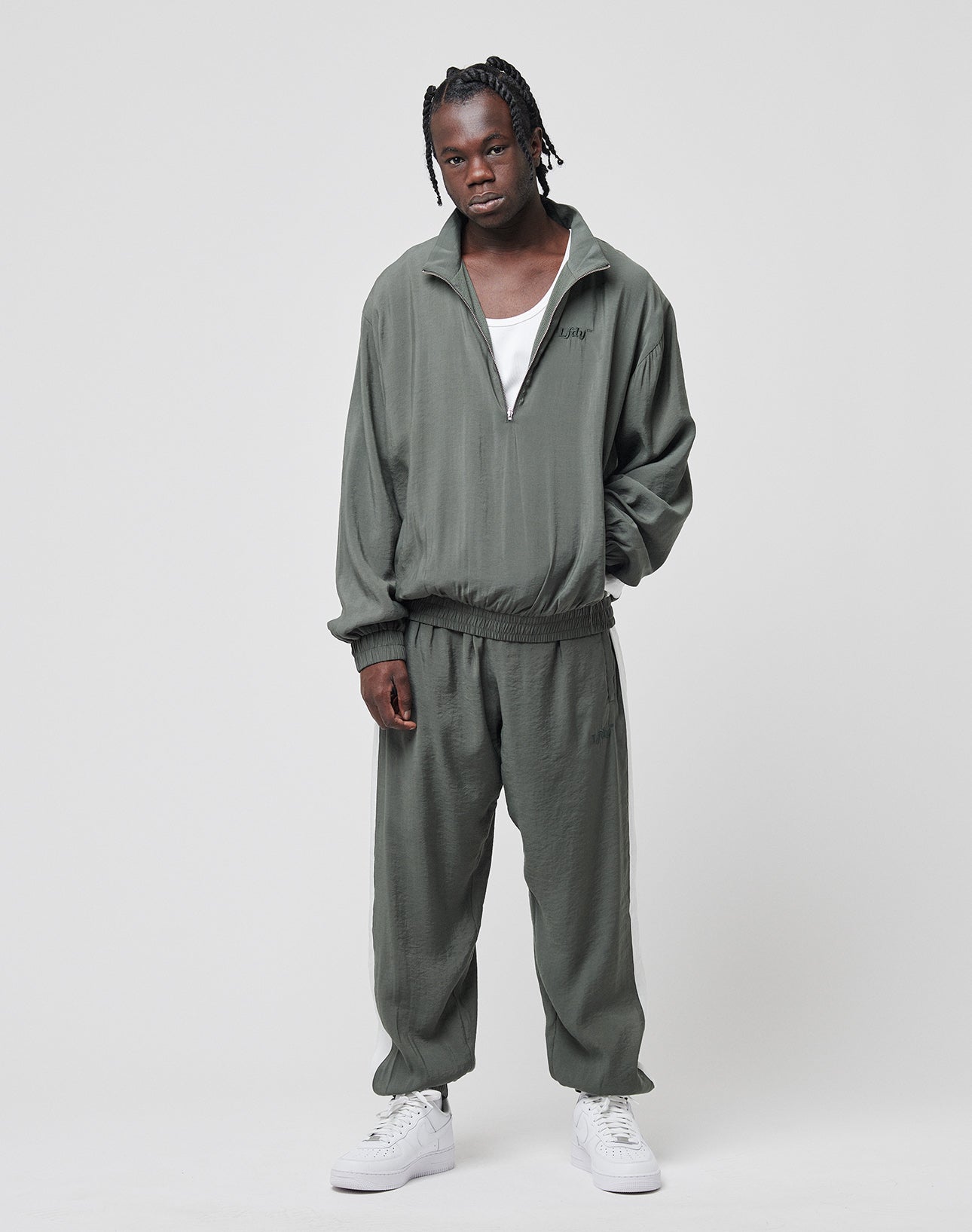 A person wearing the LIVE FAST DIE YOUNG LFDY Light Halfzip in olive green with an oversize fit and mesh lining stands facing the camera, one hand in their pocket. White sneakers, a white tank top, and braided hair complete the look against a plain background.