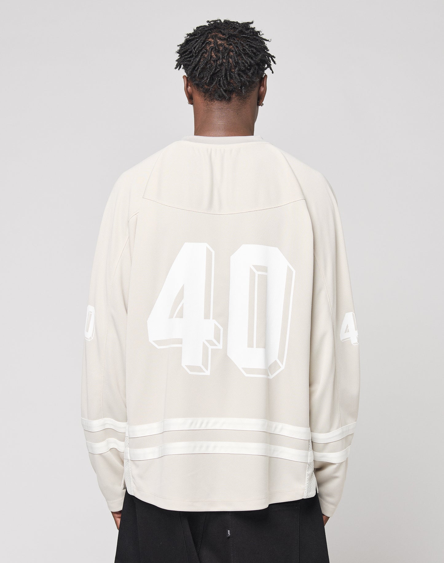 A person with short, twisted hair faces away, wearing an oversize light beige LFDY Hockey Jersey by LIVE FAST DIE YOUNG with the number 40 on the back and sleeves. The background is plain, light gray.