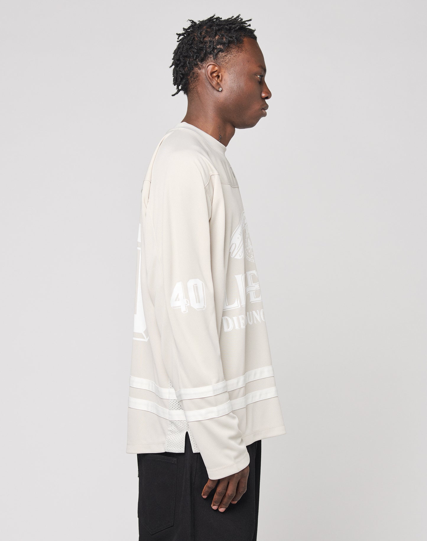 A person with short, textured hair stands in profile facing right, wearing the LIVE FAST DIE YOUNG LFDY Hockey Jersey—an oversize fit, light beige long-sleeve shirt with white graphics and Ärmelprint—paired with black pants.