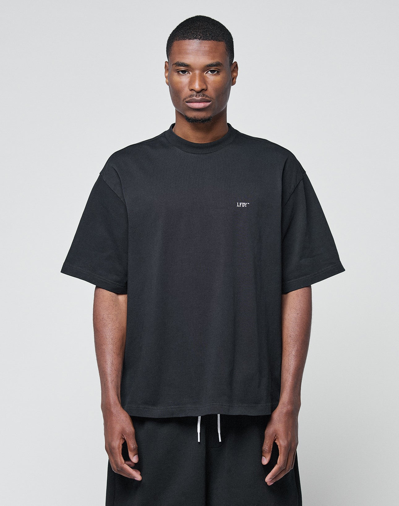 A man faces forward against a plain background, wearing the LIVE FAST DIE YOUNG Basic Trademark Tee—an oversized black 100% Baumwolle T-shirt with small white left-chest text—paired with matching black shorts.