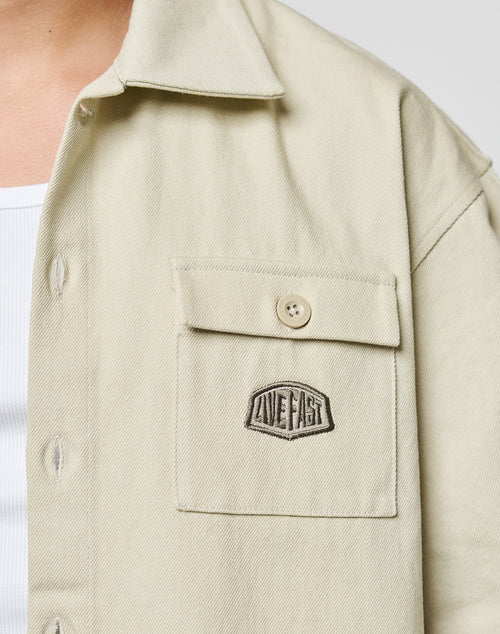 Close-up of a person wearing the LFDY Twill Shirt by LIVE FAST DIE YOUNG—a light beige cropped oversize short-sleeve made from 100% cotton, featuring a chest pocket with embroidered LIVE FAST patch, styled over a white ribbed top.