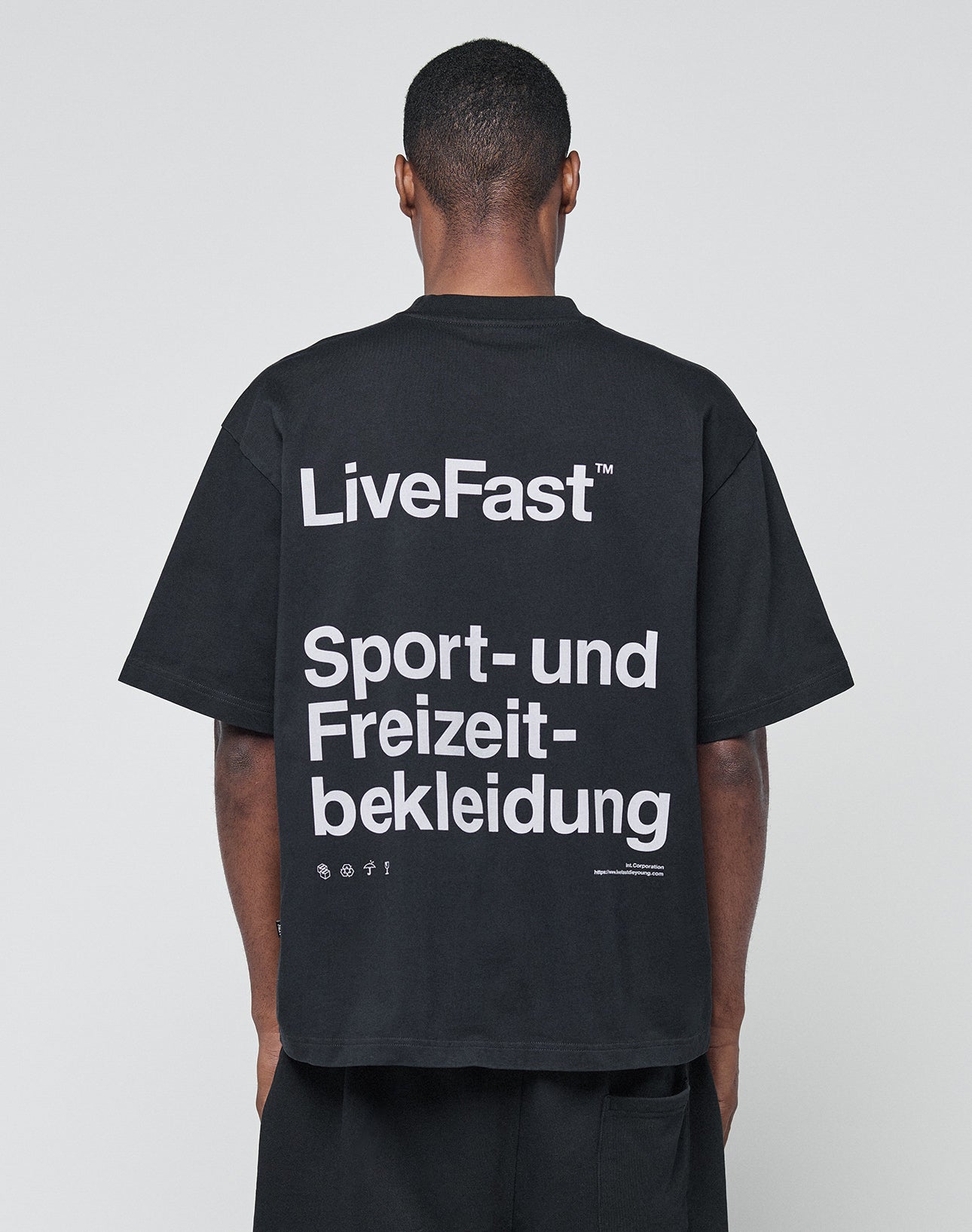 A person wearing the LIVE FAST DIE YOUNG Basic Trademark Tee, an oversize black t-shirt made from 100% Baumwolle with a bold LiveFast™ Sport- und Freizeitbekleidung backprint, faces away from the camera against a plain light background.