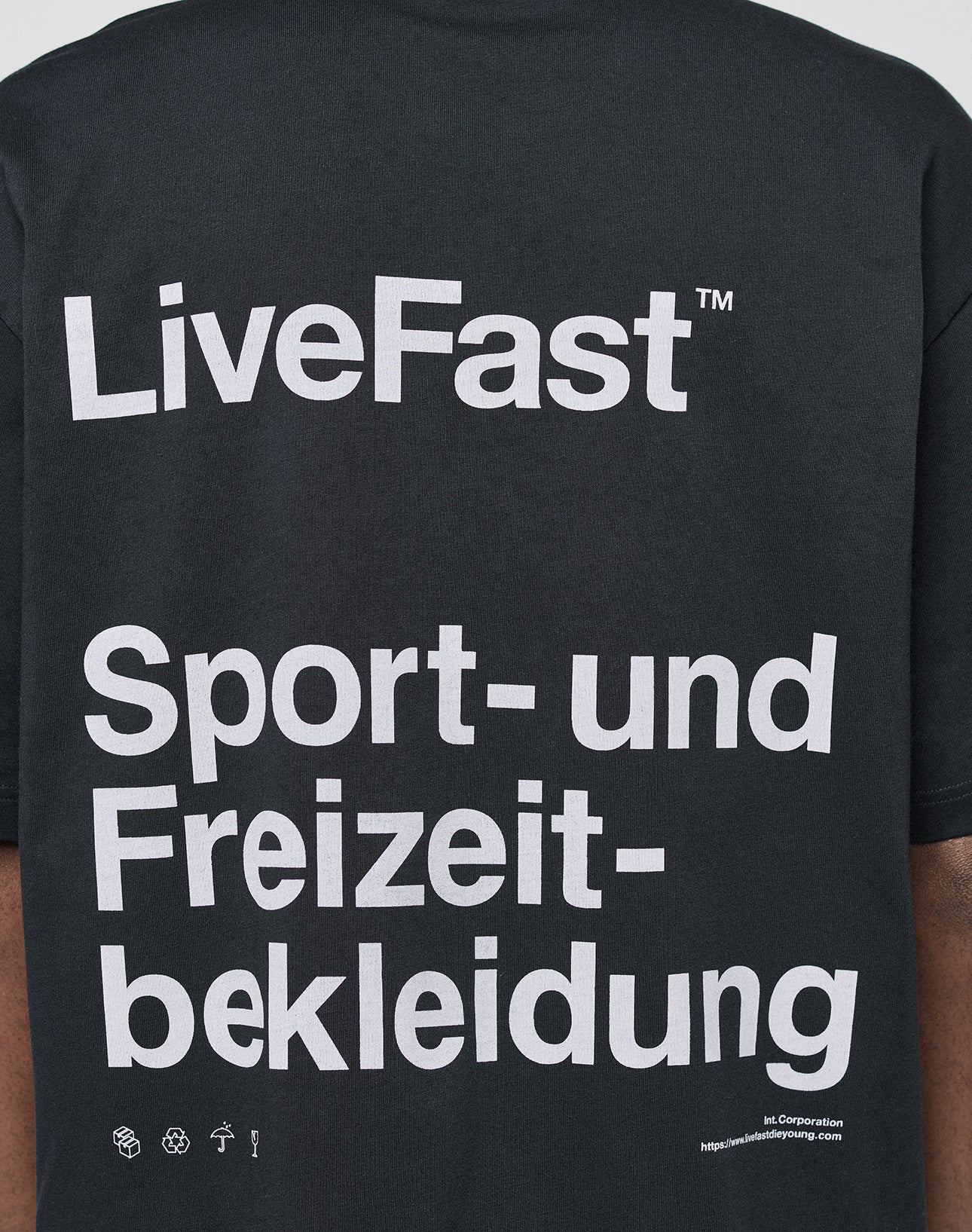 A person wears the LIVE FAST DIE YOUNG Basic Trademark Tee, a black oversized 100% cotton shirt with a white LiveFast™ Sport- und Freizeitbekleidung backprint, small icons, and website details at the bottom right. Back view shown.