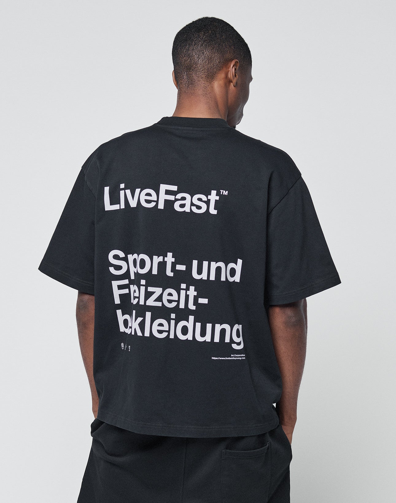 A person faces away, wearing the LIVE FAST DIE YOUNG Basic Trademark Tee—an oversize black shirt with a bold backprint: “LiveFast™ Sport- und Freizeitbekleidung.” Made from 100% Baumwolle. The background is plain and light-colored.