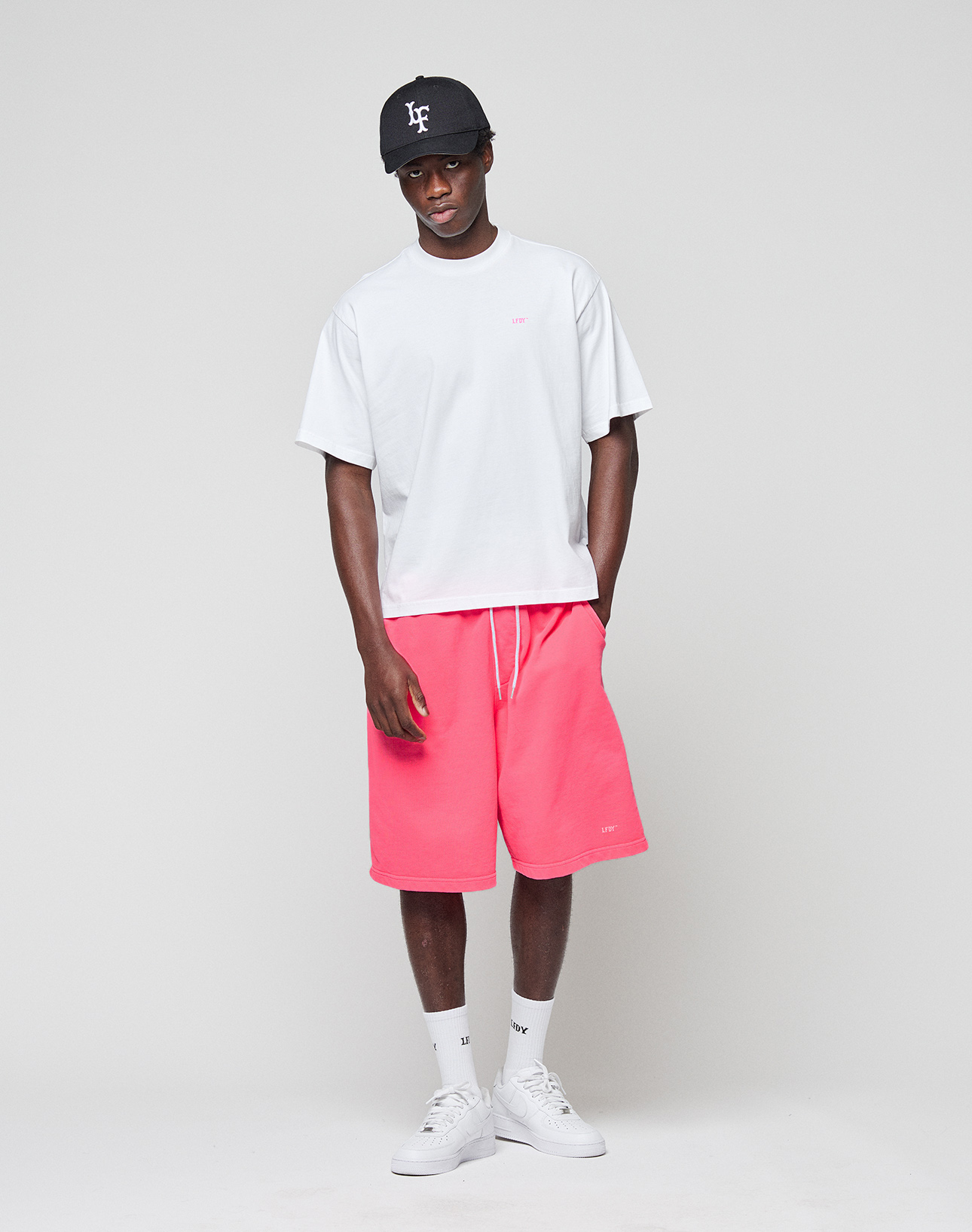 A man stands against a plain background wearing the LIVE FAST DIE YOUNG Basic Evidence Tee (100% Baumwolle, front and back print), a black cap, bright pink shorts, white crew socks, and white sneakers. He looks relaxed with his hands in his pockets.