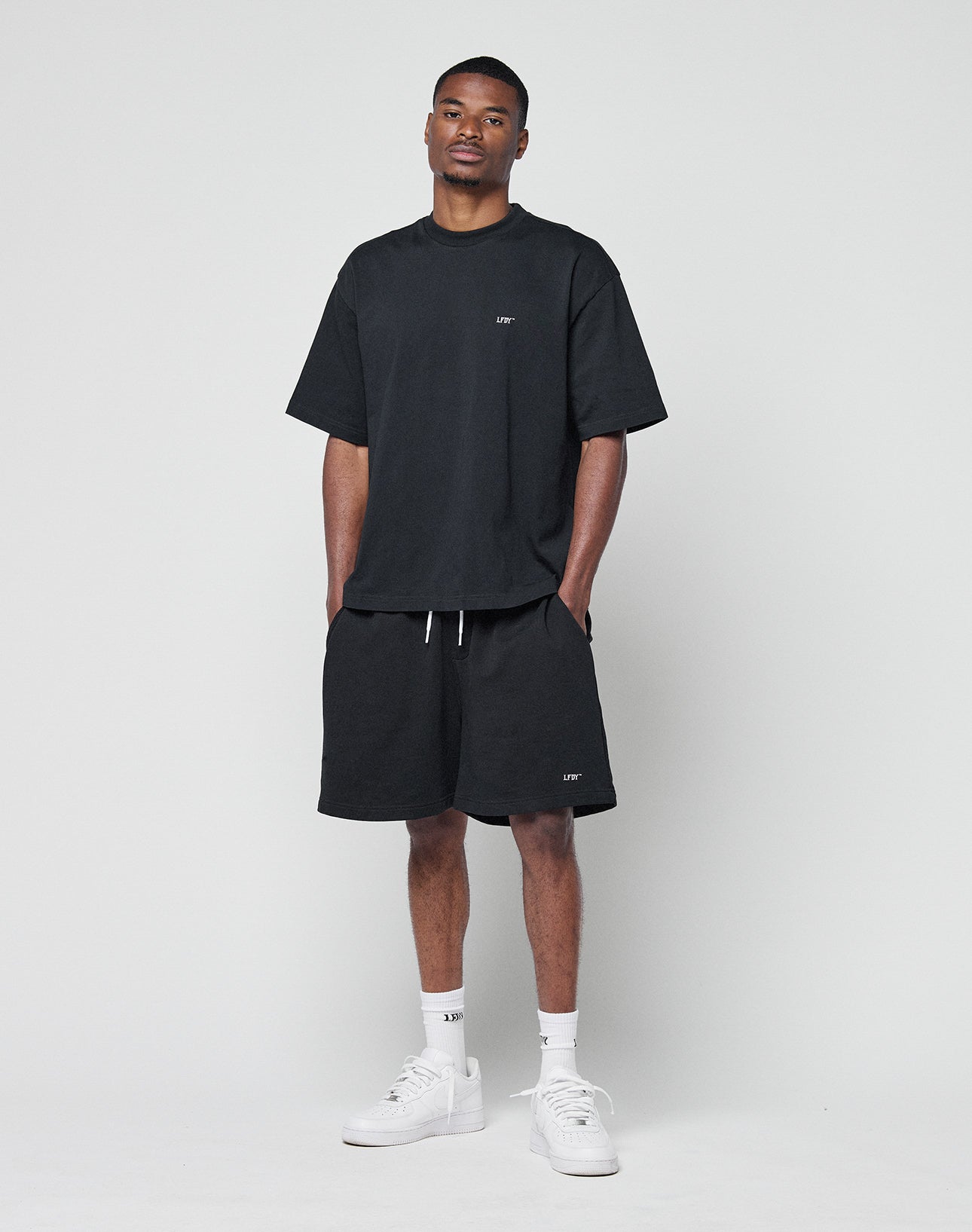 A man wears the LIVE FAST DIE YOUNG Basic Trademark Tee in black, paired with black shorts, white crew socks, and white sneakers. He stands against a plain light background with his hands in his pockets and a neutral expression.