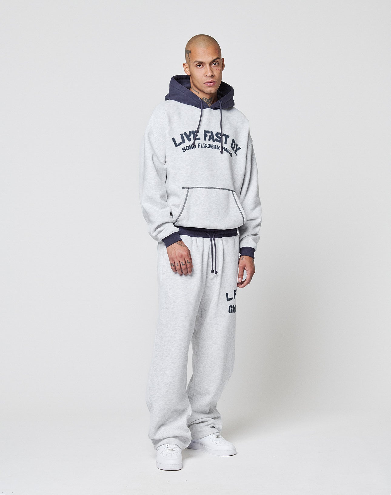 A person stands against a plain light background wearing a gray LFDY District Hooded by LIVE FAST DIE YOUNG, featuring a hood with dark contrast lining, matching gray sweatpants, and white sneakers.