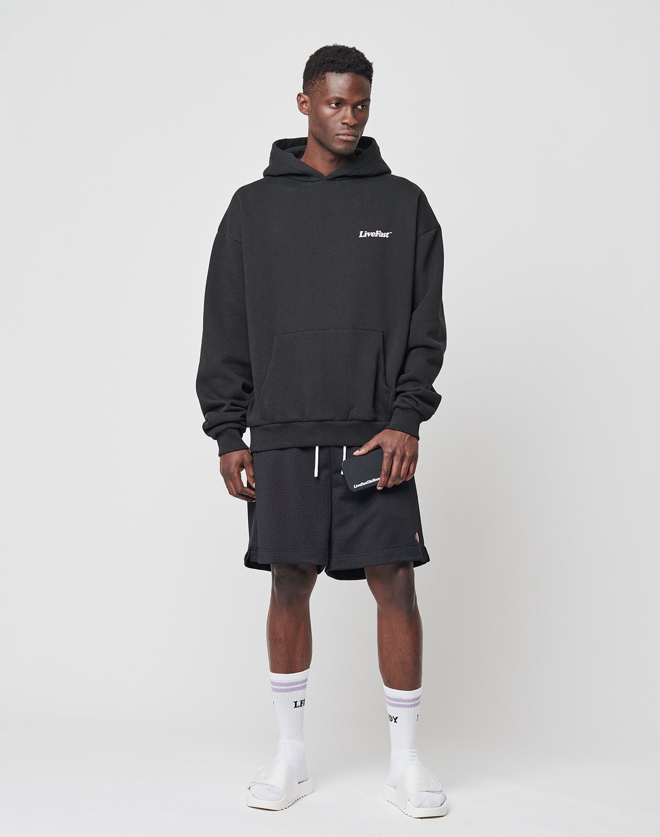 A man stands against a plain white background wearing a black hoodie, LIVE FAST DIE YOUNG Basic Mesh Shorts, white socks with LIBRARY text, slide sandals, and a wallet. A logo patch adds detail to his look.