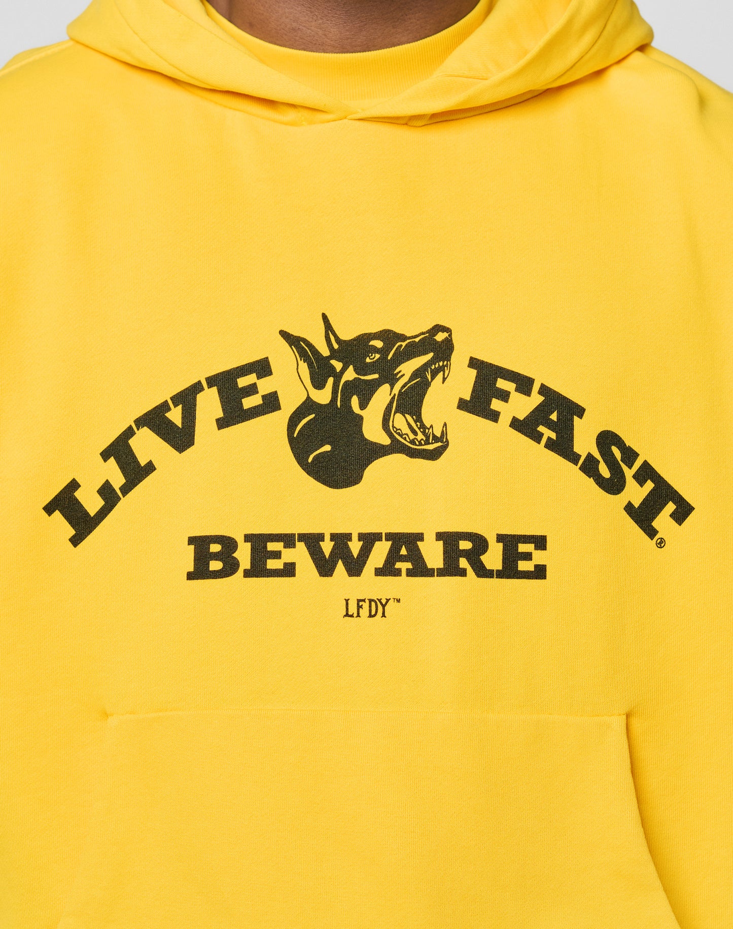 The Live Fast Beware Hooded by LIVE FAST DIE YOUNG is a yellow oversized cotton hoodie with a black snarling dog graphic, LIVE FAST above, and BEWARE plus LFDY below.