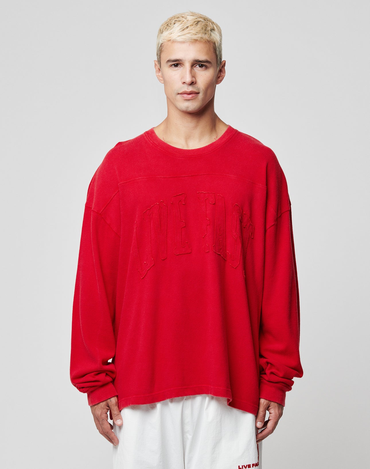 A person with short blonde hair wears the LIVE FAST DIE YOUNG Ripped College Longsleeveāan oversize red sweatshirt with subtle textāand loose white shorts, standing against a plain light gray background.
