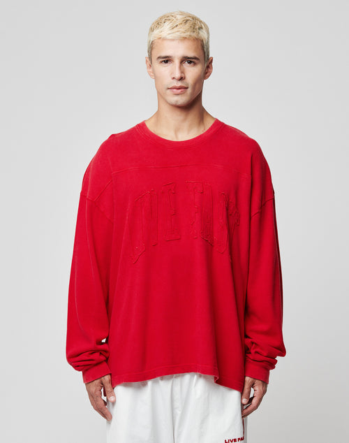 A person with short blonde hair wears the LIVE FAST DIE YOUNG Ripped College Longsleeve—an oversize red sweatshirt with subtle text—and loose white shorts, standing against a plain light gray background.