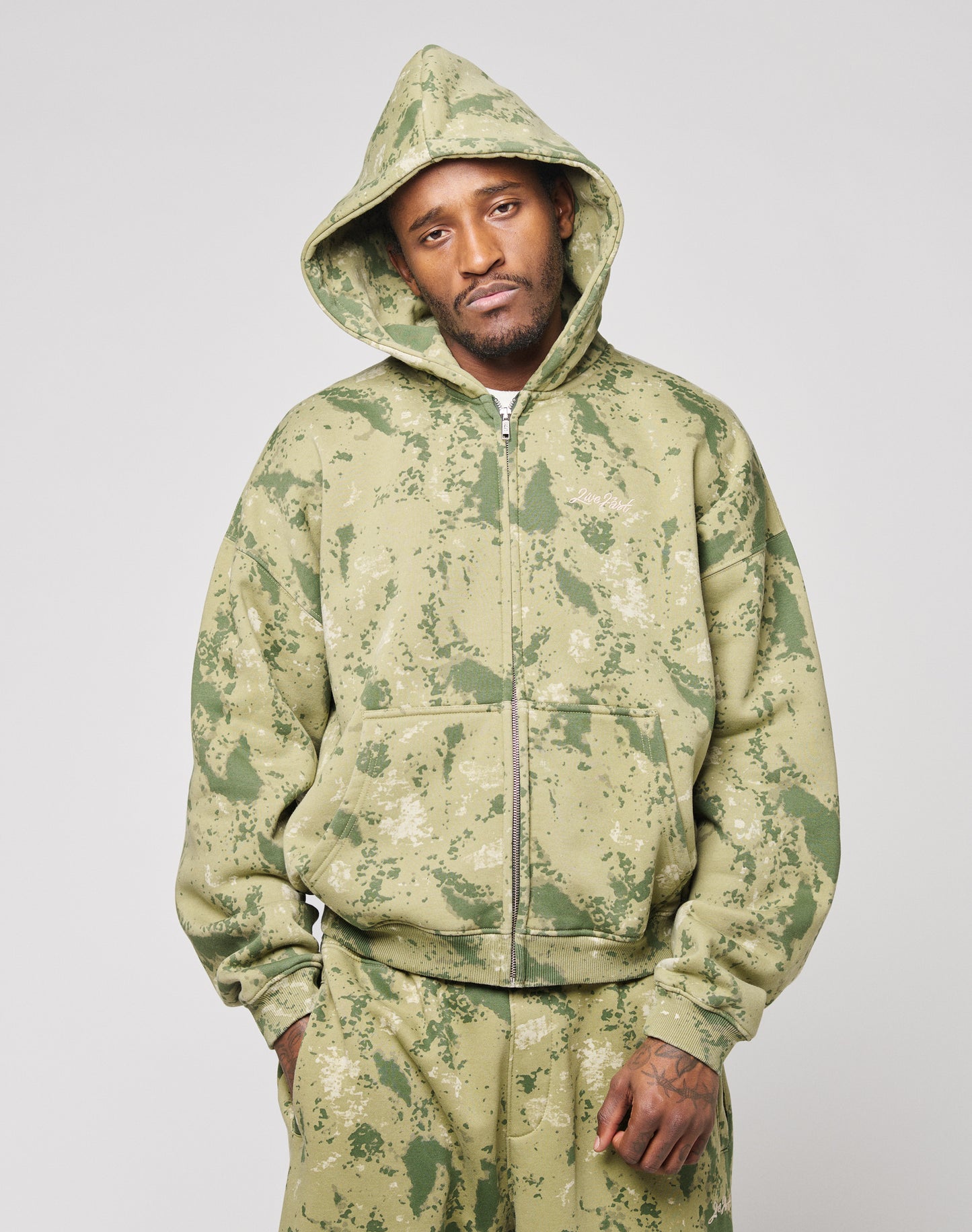 A man wears the LIVE FAST DIE YOUNG Camo Hooded Zipāa green and beige oversized camo-patterned hoodieāpaired with matching pants, standing against a plain background and looking at the camera with a neutral expression.