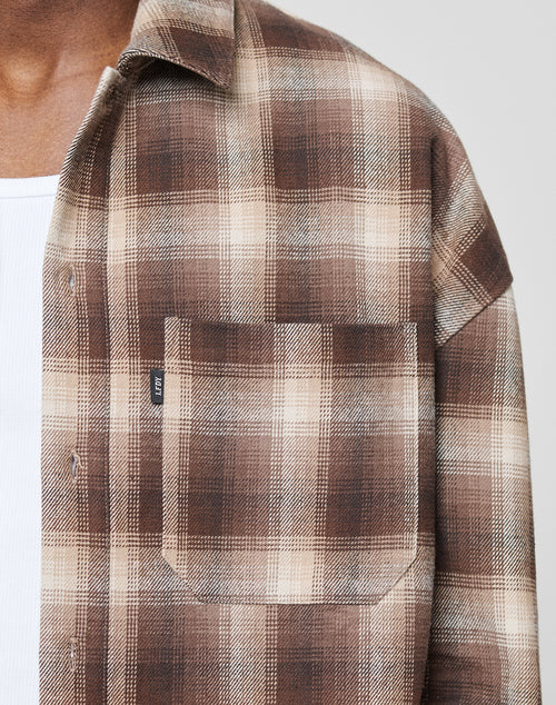 A close-up shows a person wearing the LIVE FAST DIE YOUNG Vintage Flannel in brown and beige plaid over a white ribbed undershirt. Only the chest and neck are visible, with an LFDY-branded front pocket on a small black tag.