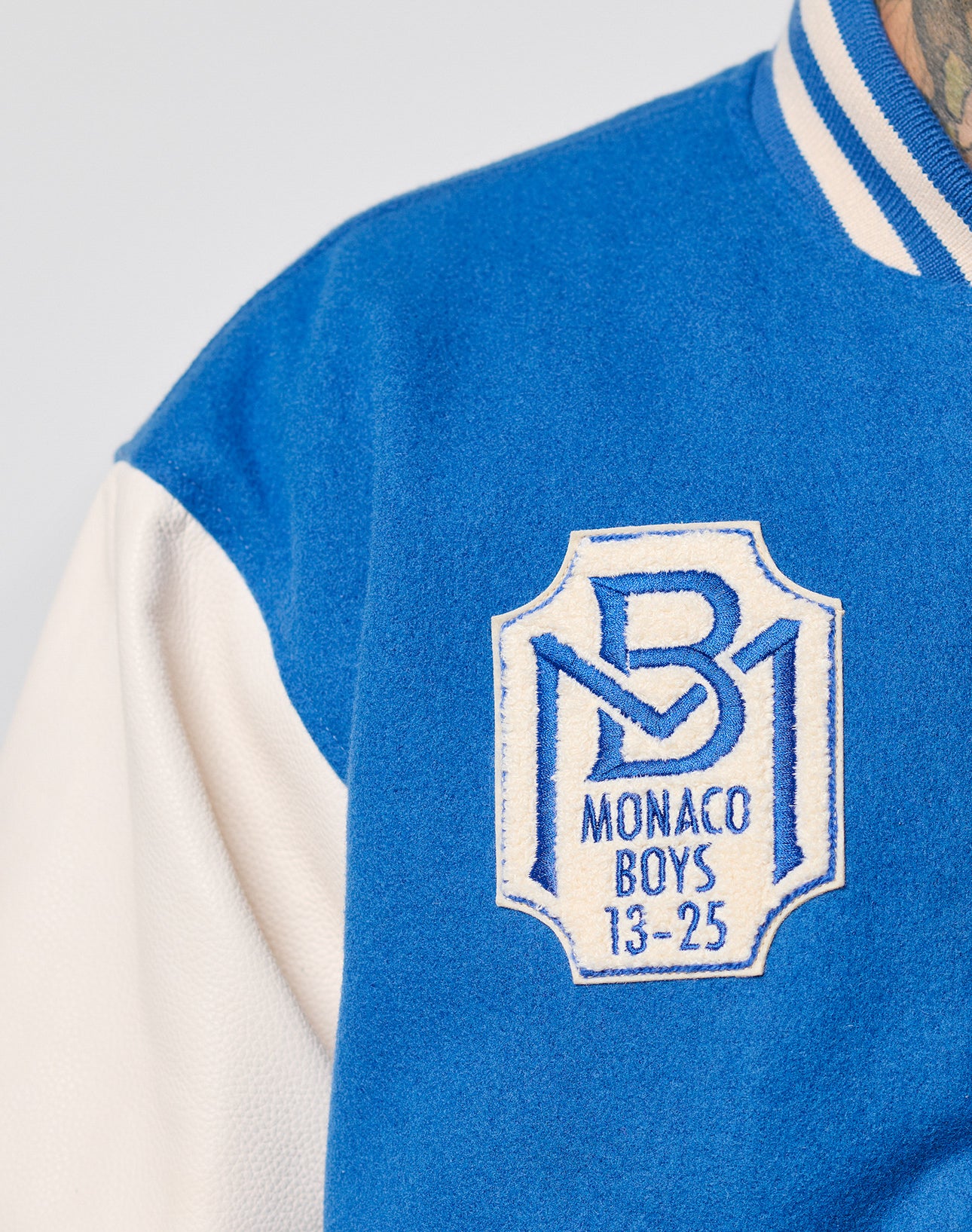 Close-up of the LIVE FAST DIE YOUNG x Schott NYC Varsity Jacket in blue and white, featuring cowhide leather sleeves and a chenille Monaco Boys 13-25 patch with a stylized MB logo on the chest.