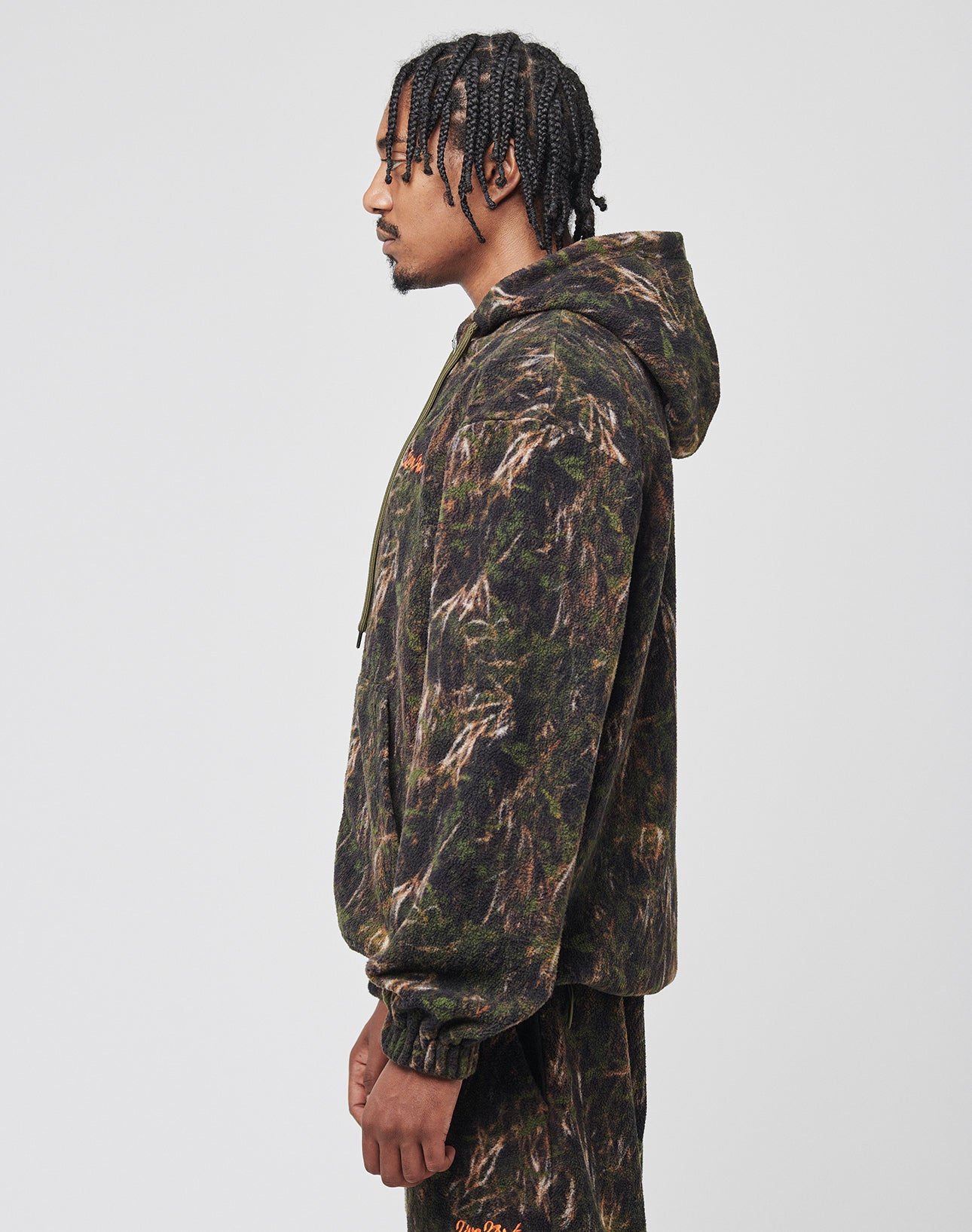 A man with braided hair stands in profile, wearing a LIVE FAST DIE YOUNG LFDY Fleece Hooded Zip and matching green and brown camouflage pants, set against a plain light background.