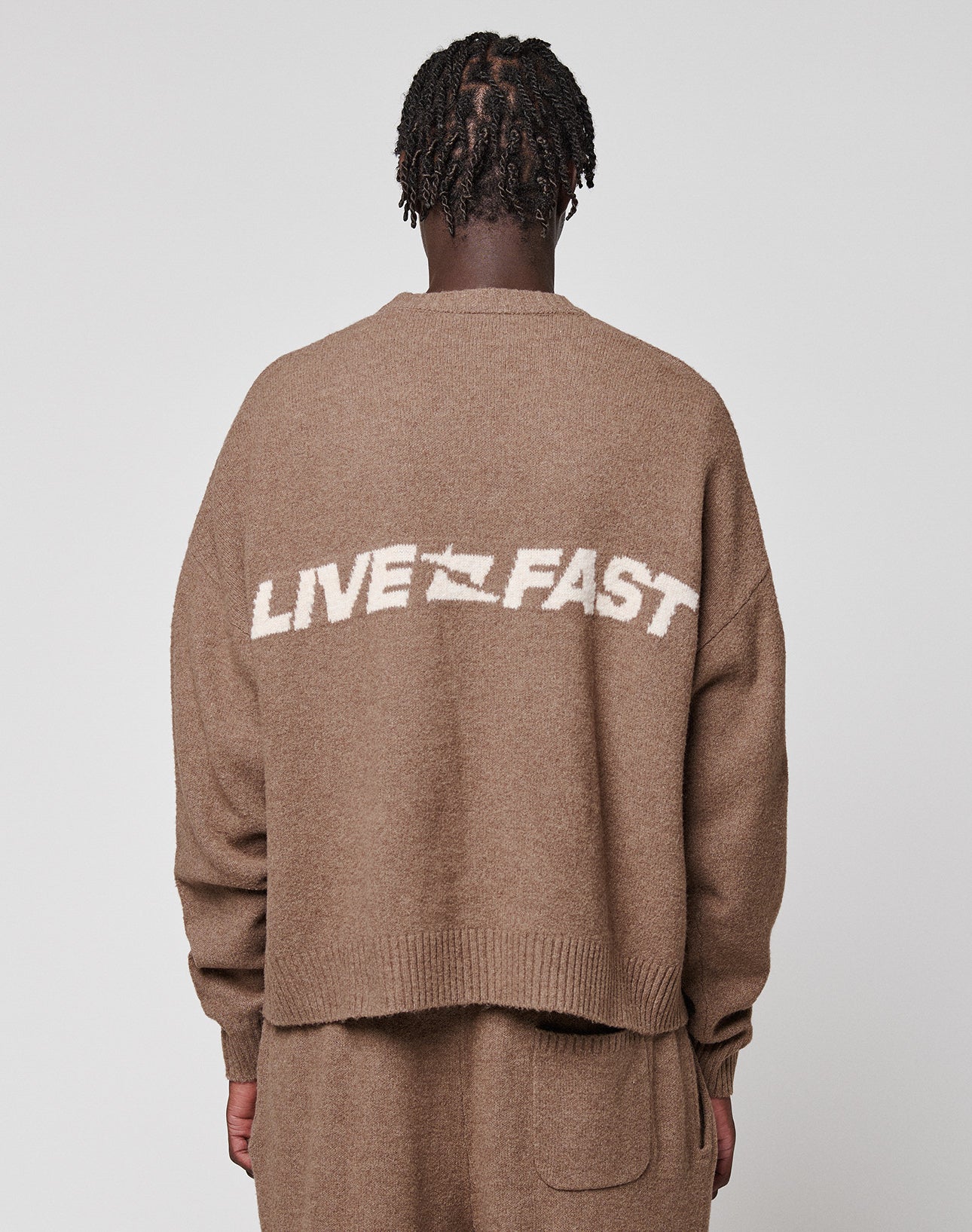 Seen from behind, a person with short, twisted hair wears the LIVE FAST DIE YOUNG Live Fast Boucle Sweater in an oversize fit brown, featuring "LIVE FAST" in white and a streaked Strick design on the back. Matching pants complete the look.