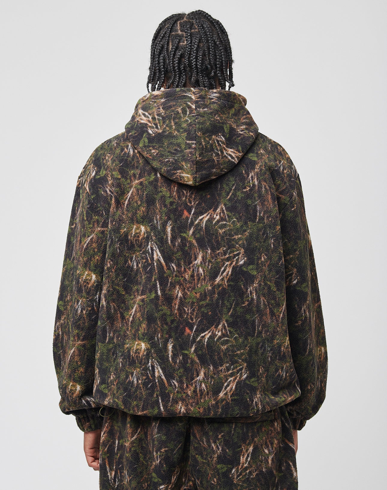 A person with braided hair, seen from behind, wears the LFDY Fleece Hooded Zip by LIVE FAST DIE YOUNG and matching camouflage pants in green and brown hues. The Oversize Fit gives a relaxed vibe against the light background.