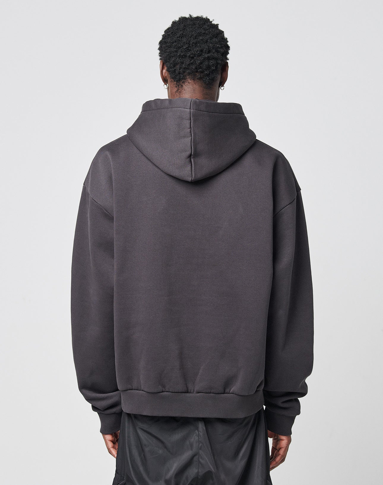 A person faces away from the camera, wearing the LIVE FAST DIE YOUNG Its Vulgar Center Hooded in dark gray with hood down and loose black pants, set against a plain light background.