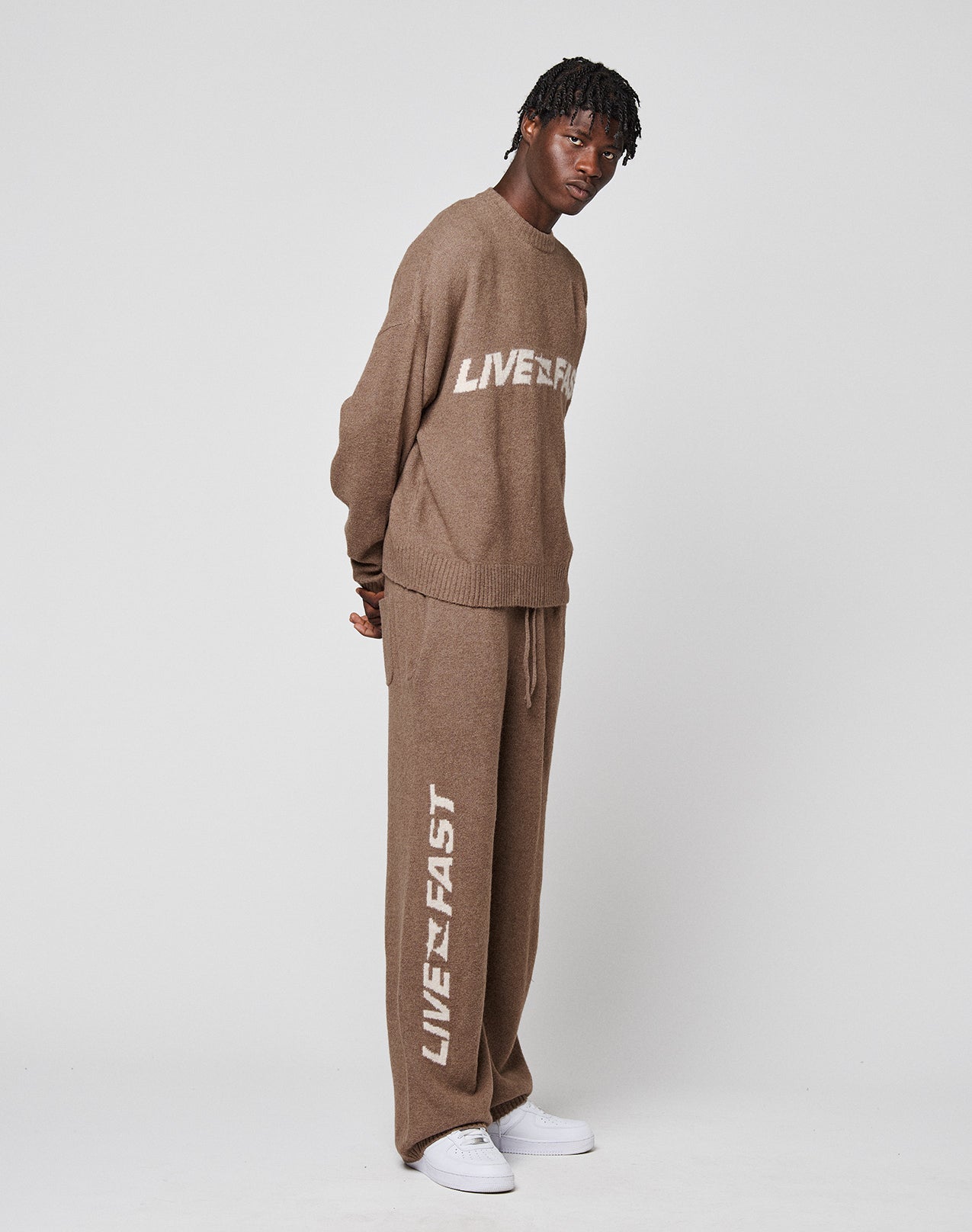 A person poses against a plain background in an oversize fit Live Fast Boucle Sweater by LIVE FAST DIE YOUNG and matching pants, both brown with LIVEFAST in white, styled with white sneakers.