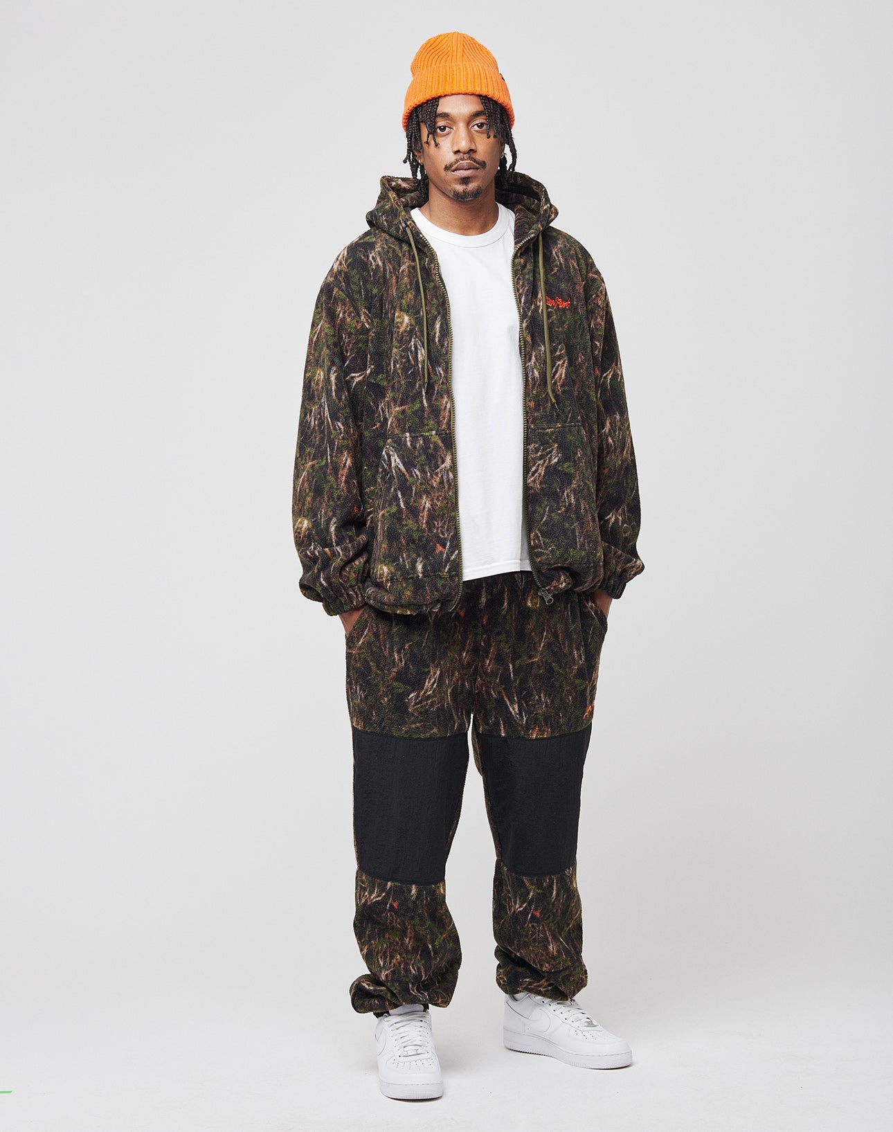 A man in an LFDY Fleece Hooded Zip by LIVE FAST DIE YOUNG, paired with a white T-shirt, orange beanie, and white sneakers, stands against a plain light gray background.