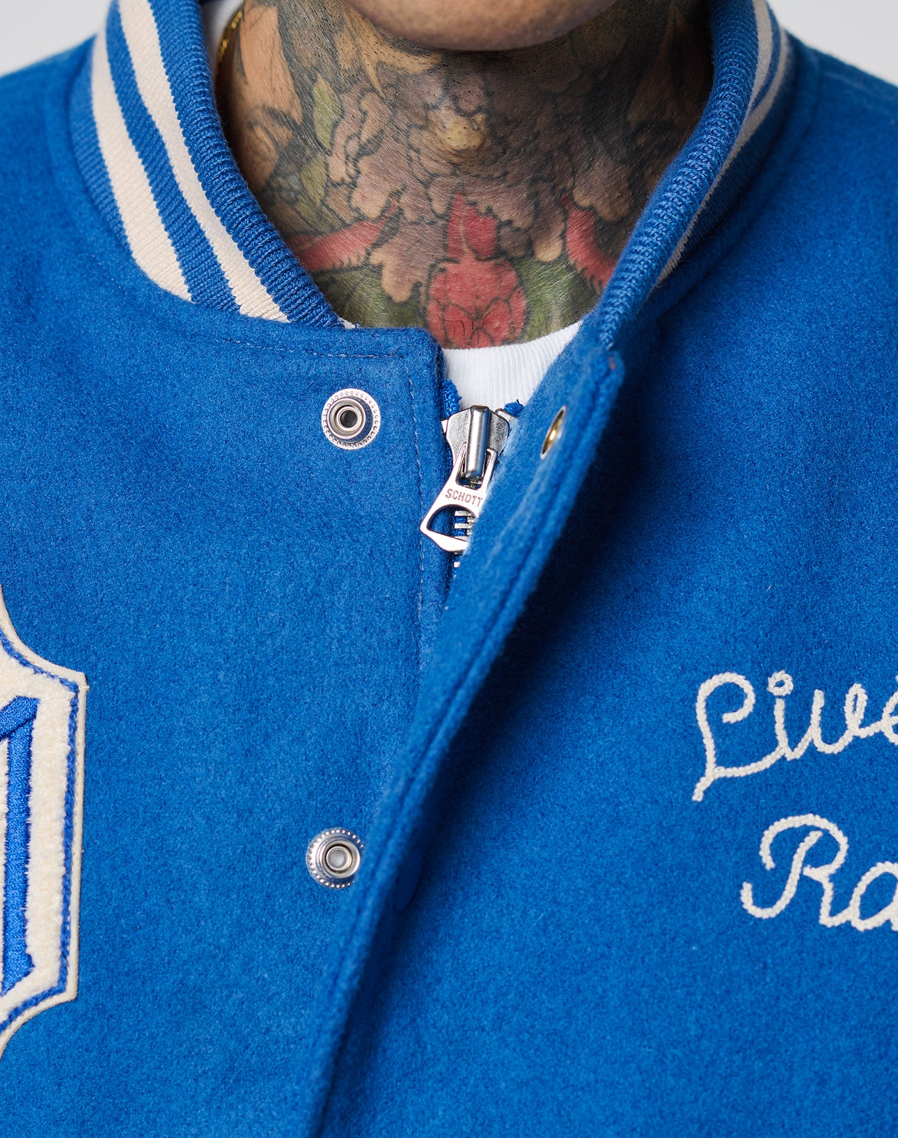 Close-up of a person in a LIVE FAST DIE YOUNG LFDY x Schott NYC Varsity Jacket with chenille patches, cowhide leather sleeves, white trim, and embroidered text. Colorful floral neck tattoos are visible above a white shirt.