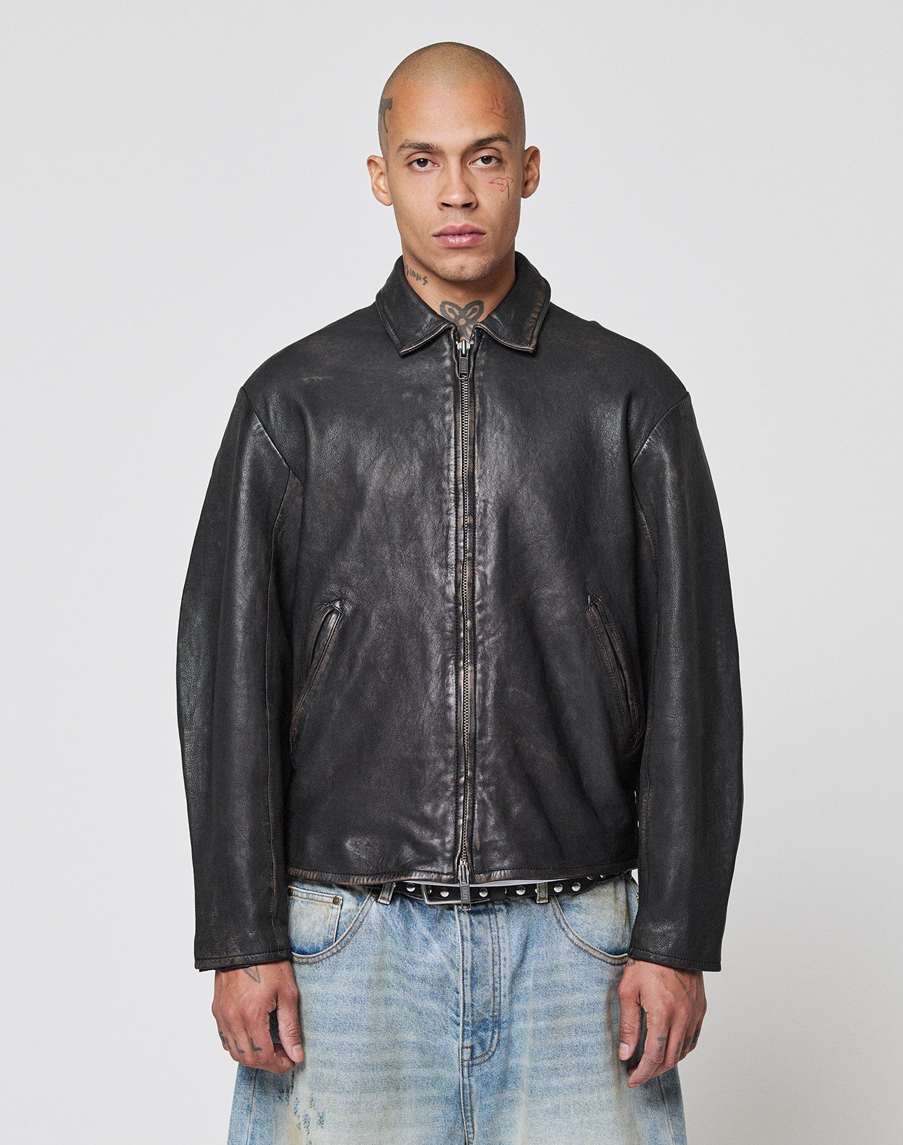 A person with a shaved head wears a black LIVE FAST DIE YOUNG LFDY Leather Jacket with zipper and light blue jeans, standing facing forward against a plain white background.