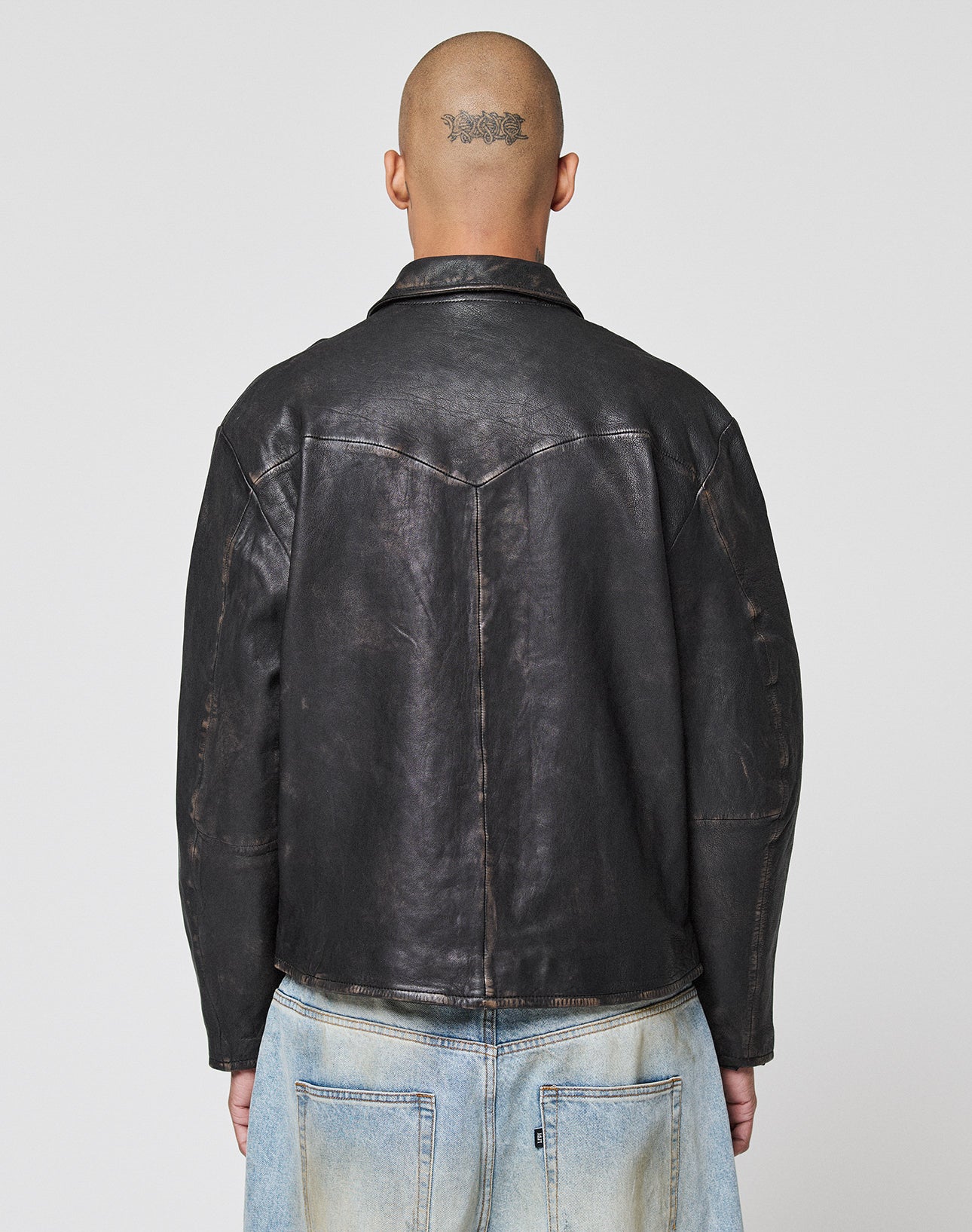 A person with a shaved head and a tattoo on the back faces away from the camera, wearing a black LFDY Leather Jacket by LIVE FAST DIE YOUNG and light blue denim jeans against a plain background.