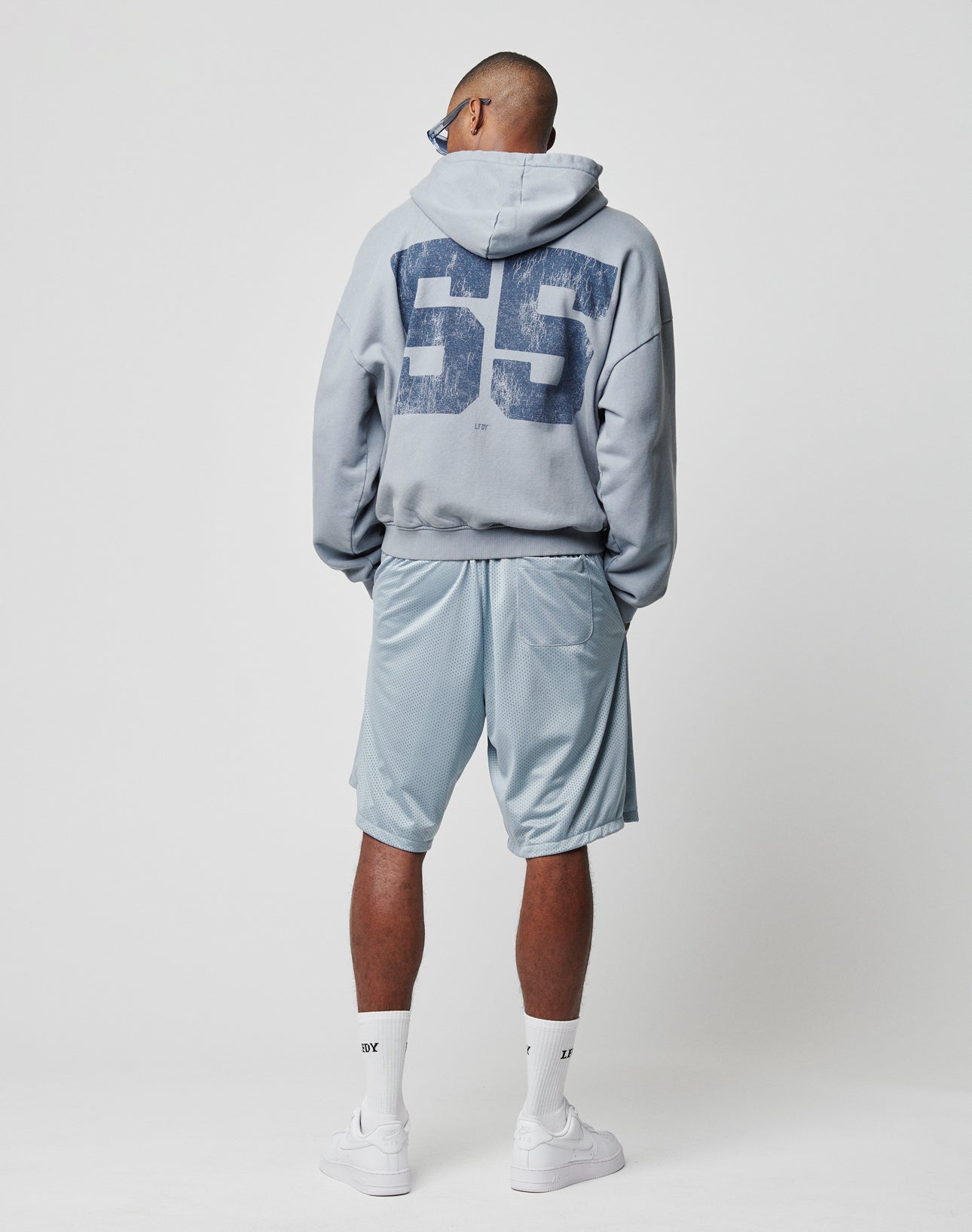A person stands with their back turned, wearing the LIVE FAST DIE YOUNG LFDY 55 Hooded Zip in light blue with 55 on the back, matching vintage wash shorts, white socks, and white sneakers against a plain white background.