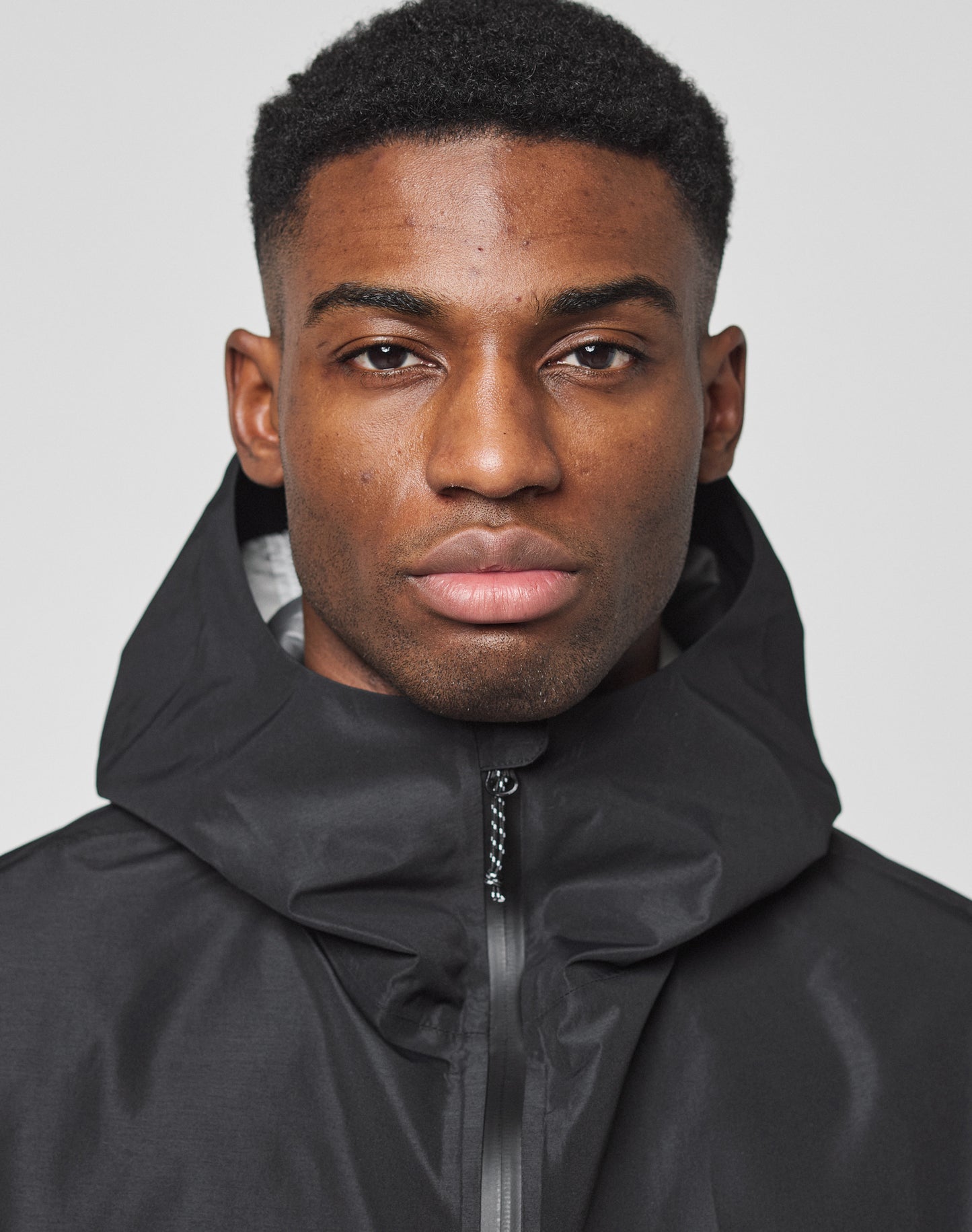 A man with short black hair wears the LIVE FAST DIE YOUNG Live Fast Performance Rain Jacket, featuring wasserdichte Nähte, zipped to the top, and looks neutrally at the camera against a plain light background.