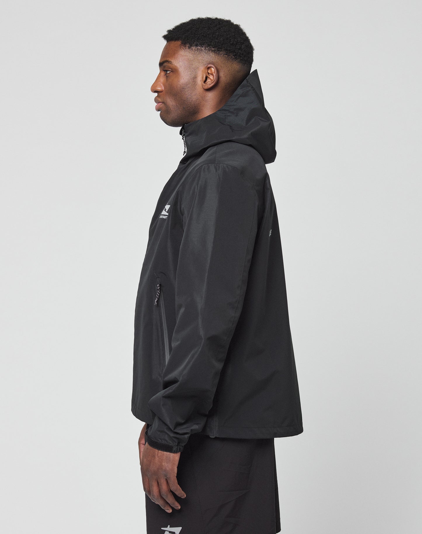 A man in a black LIVE FAST DIE YOUNG Live Fast Performance Rain Jacket with visor hood and black shorts stands in profile against a plain light gray background.