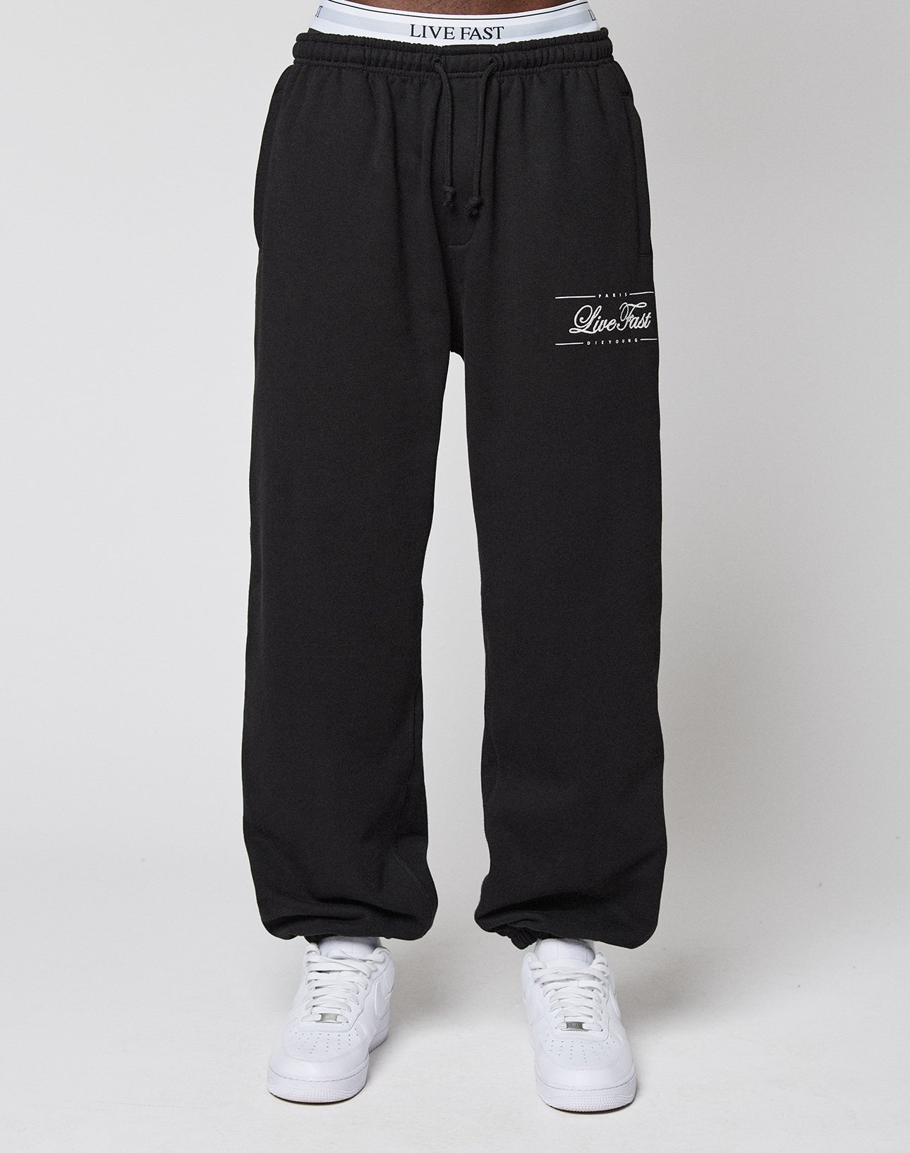 A person wearing black Wide Cuff Jogger sweatpants by LIVE FAST DIE YOUNG, featuring a Live Fast logo on the left thigh, a comfortable elastic waistband, soft French Terry fabric, and paired with white sneakers against a plain white background.