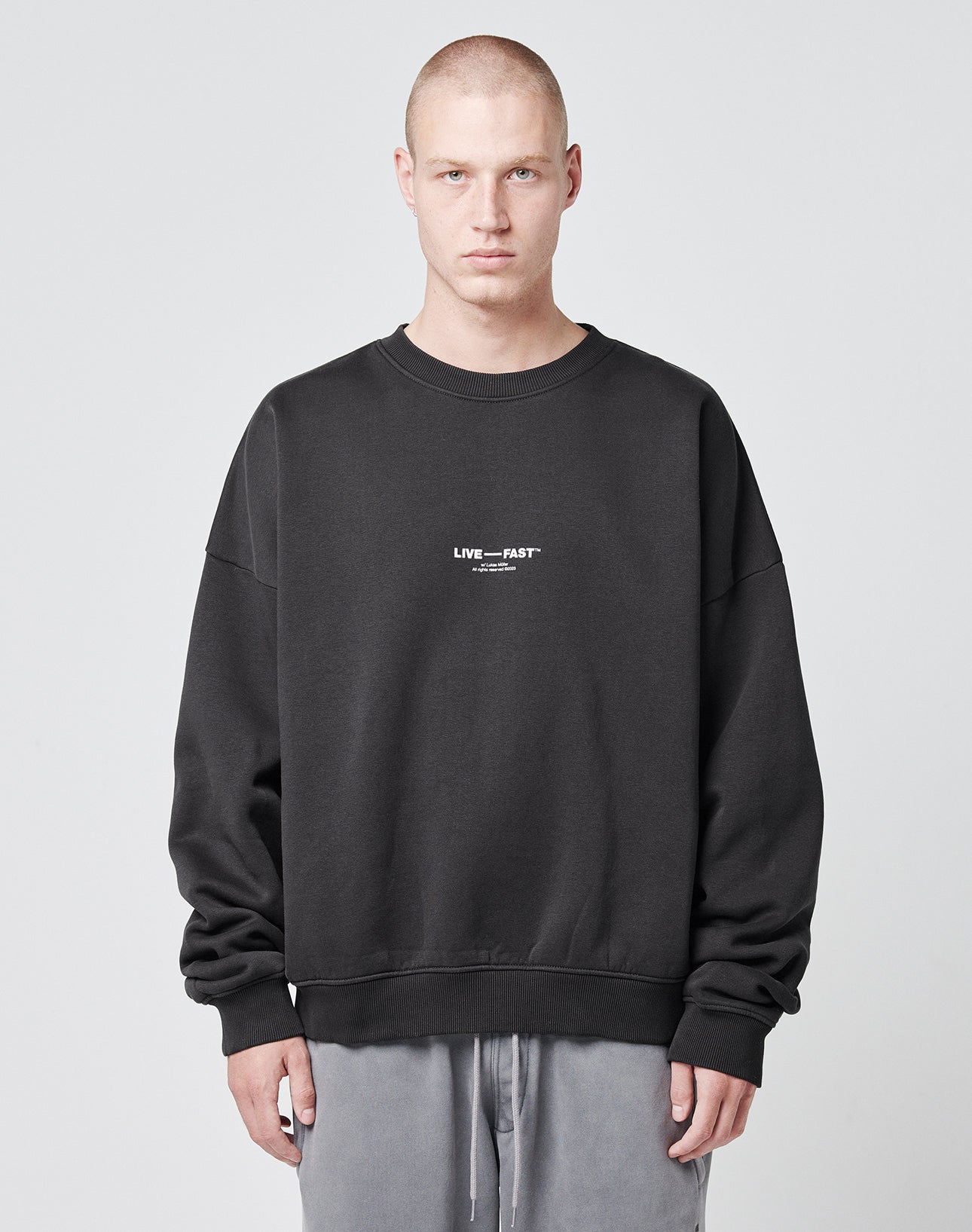 A person with a shaved head models the Tape Sweater by LIVE FAST DIE YOUNGāan oversized black sweatshirt with LIVE FAST in small white text on the chestāpaired with light gray pants against a plain light gray background.