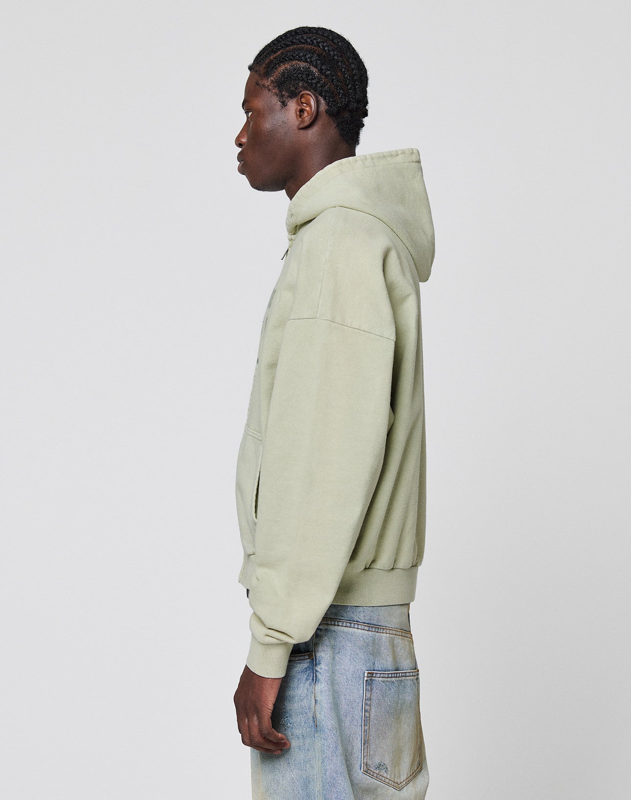 A person stands in profile against a plain background, wearing the LIVE FAST DIE YOUNG LFDY District Hooded Zip in light green with an Oversize Fit and light blue jeans.